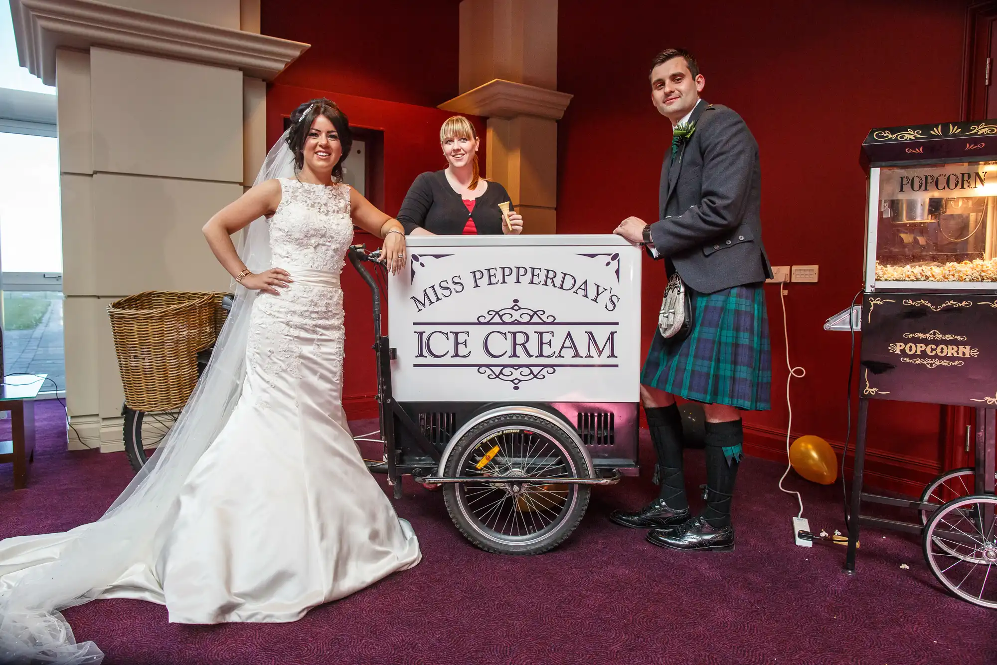 A bride and groom stand with a woman serving ice cream from a vintage cart at a wedding reception.