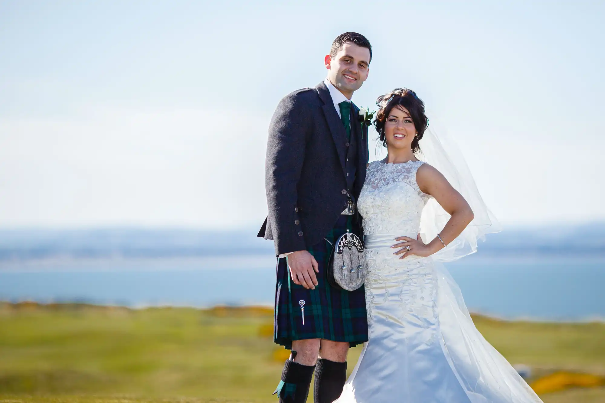 A bride and groom smiling outdoors, the groom in a kilt, with a scenic coastal backdrop.