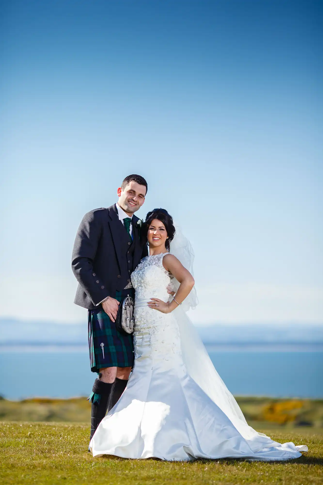 A bride in a white dress and a groom in a kilt stand together on a grassy field with a clear blue sky and distant sea behind them.