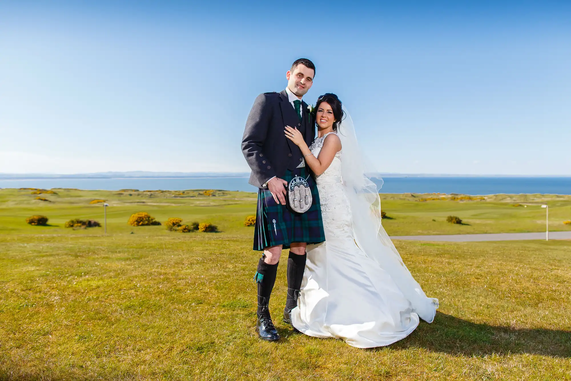 A bride in a white gown and a groom in a traditional scottish kilt posing on a grassy field with the ocean and a clear sky in the background.