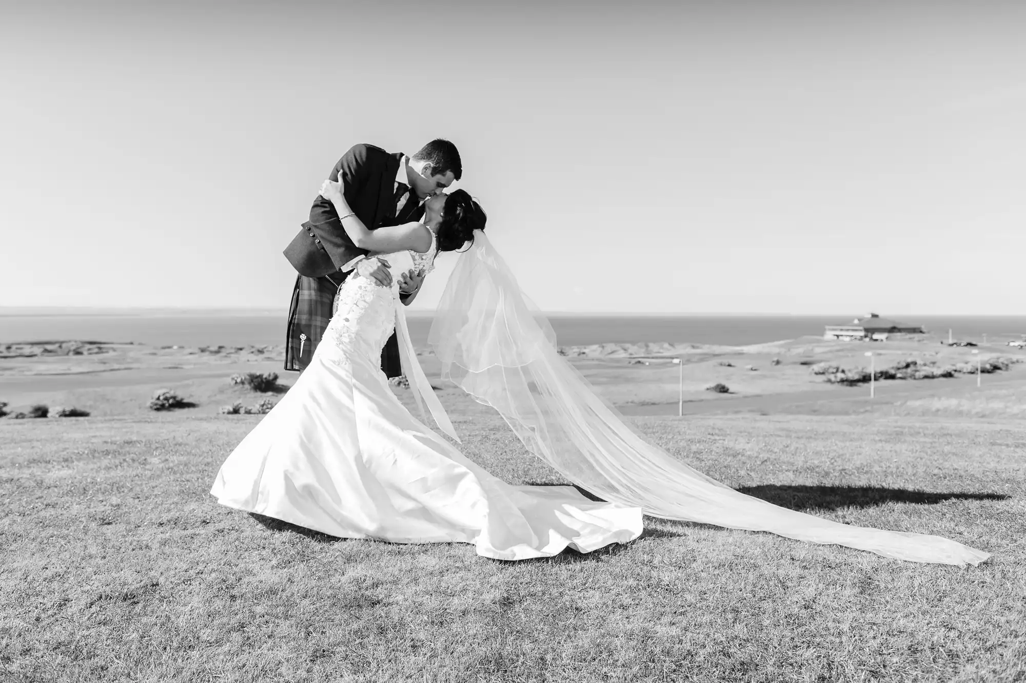 A bride and groom share a kiss on a grassy seaside meadow; the groom wears a kilt, and the bride's long veil flows elegantly in the breeze, with a coastal landscape in the background.