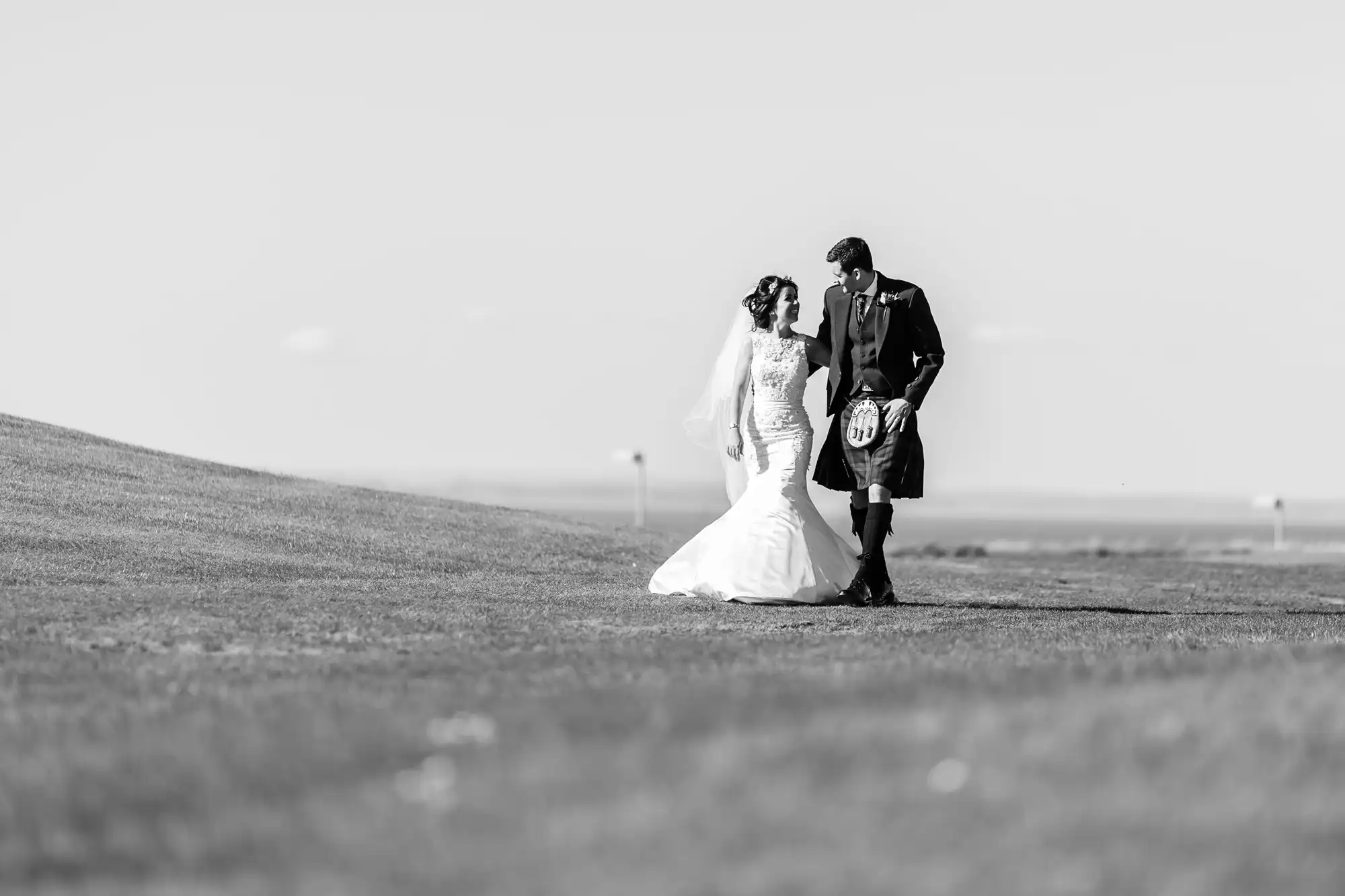 A bride and groom in formal wedding attire hold hands on a vast, grassy field under a clear sky, standing together in a serene black and white setting.