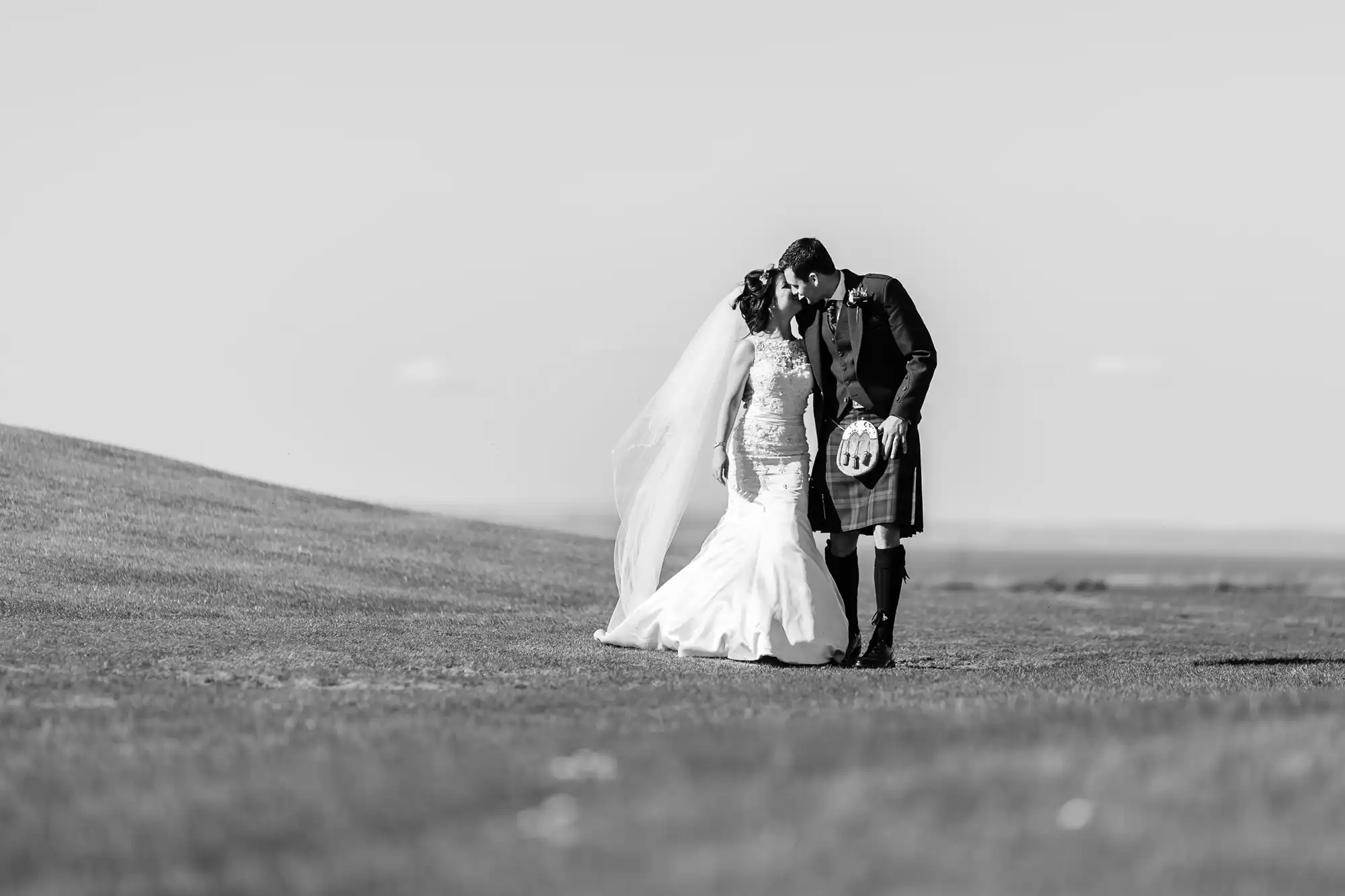 A bride and groom in wedding attire embrace tenderly in a vast, open field, with the groom dressed in a traditional kilt. the image is in black and white.