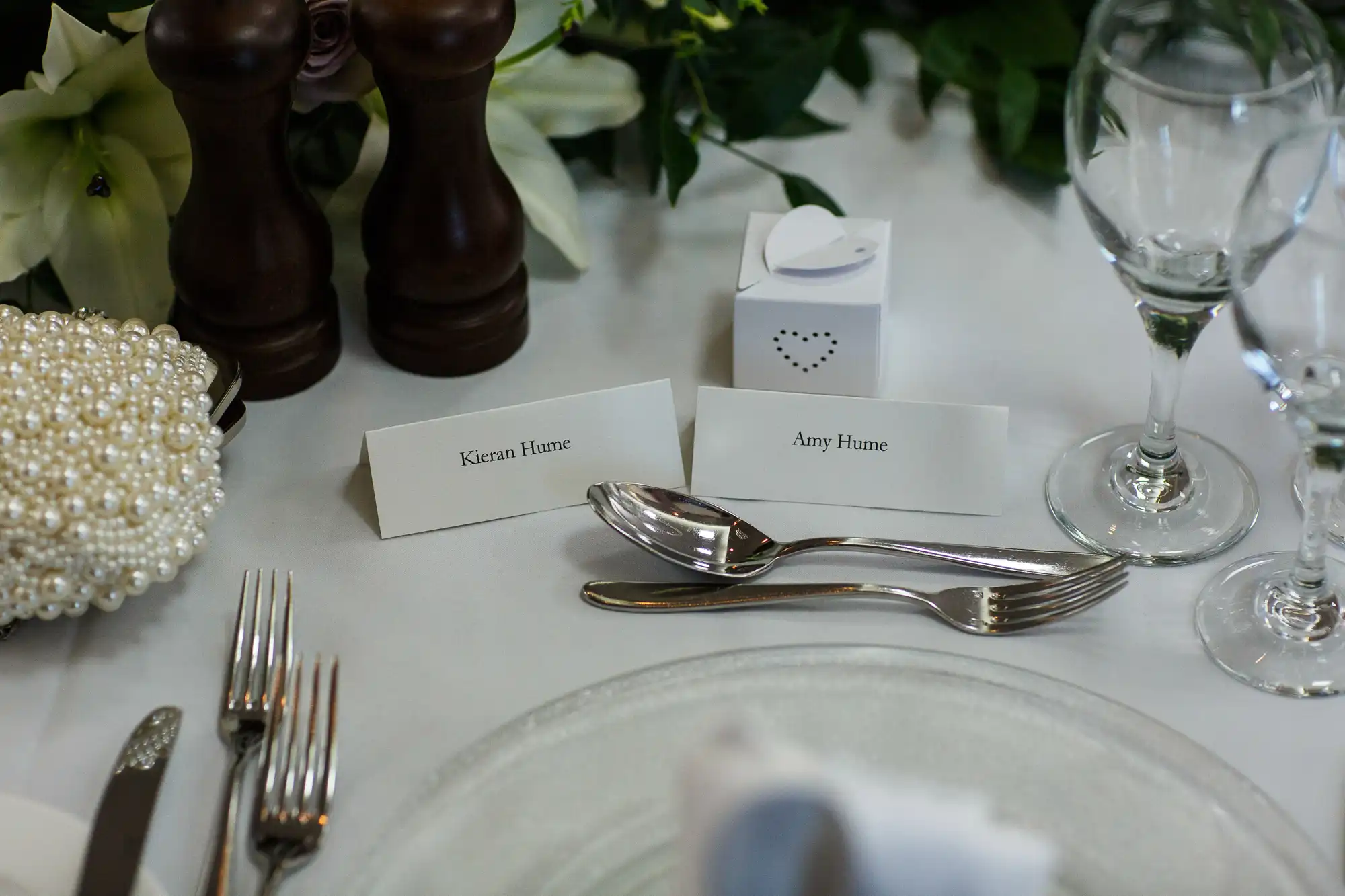 Elegant wedding table setting with name cards, pearl necklace, silver cutlery, and ivy decoration on a white tablecloth.