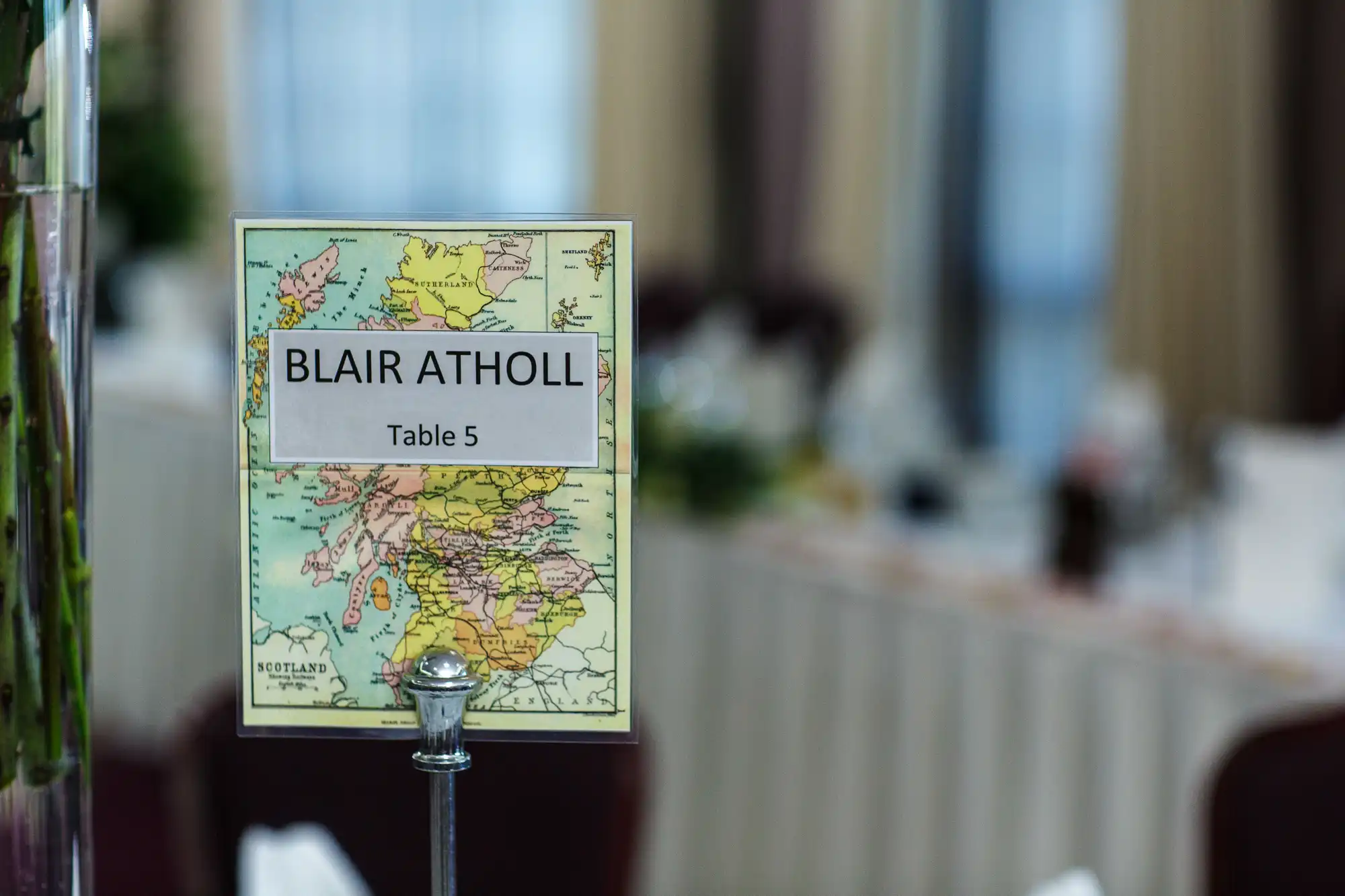A table marker with "blair atholl - table 5" over a colorful map, with a blurred background of a banquet hall.