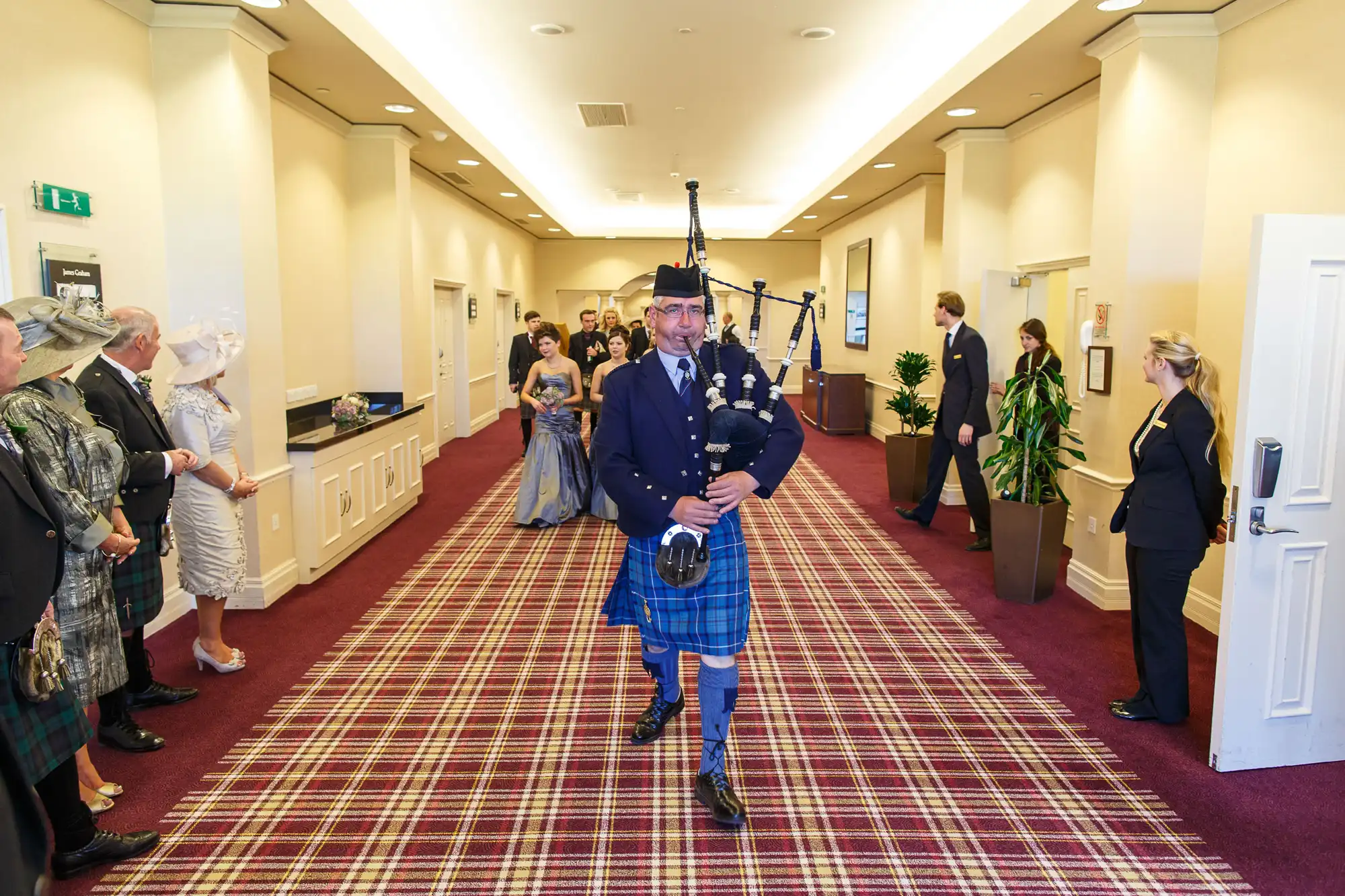 A bagpiper in traditional scottish attire playing in a hotel corridor, with spectators in formal wear observing.