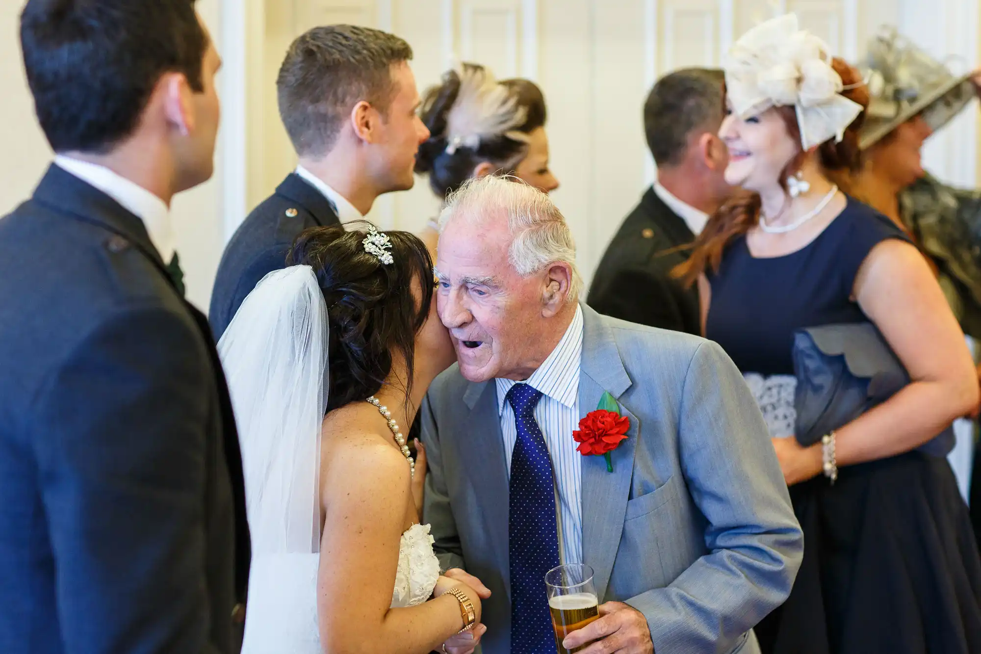 Elderly man in a suit with a red flower pin sharing a joyful moment with a bride at a wedding reception, surrounded by other guests.