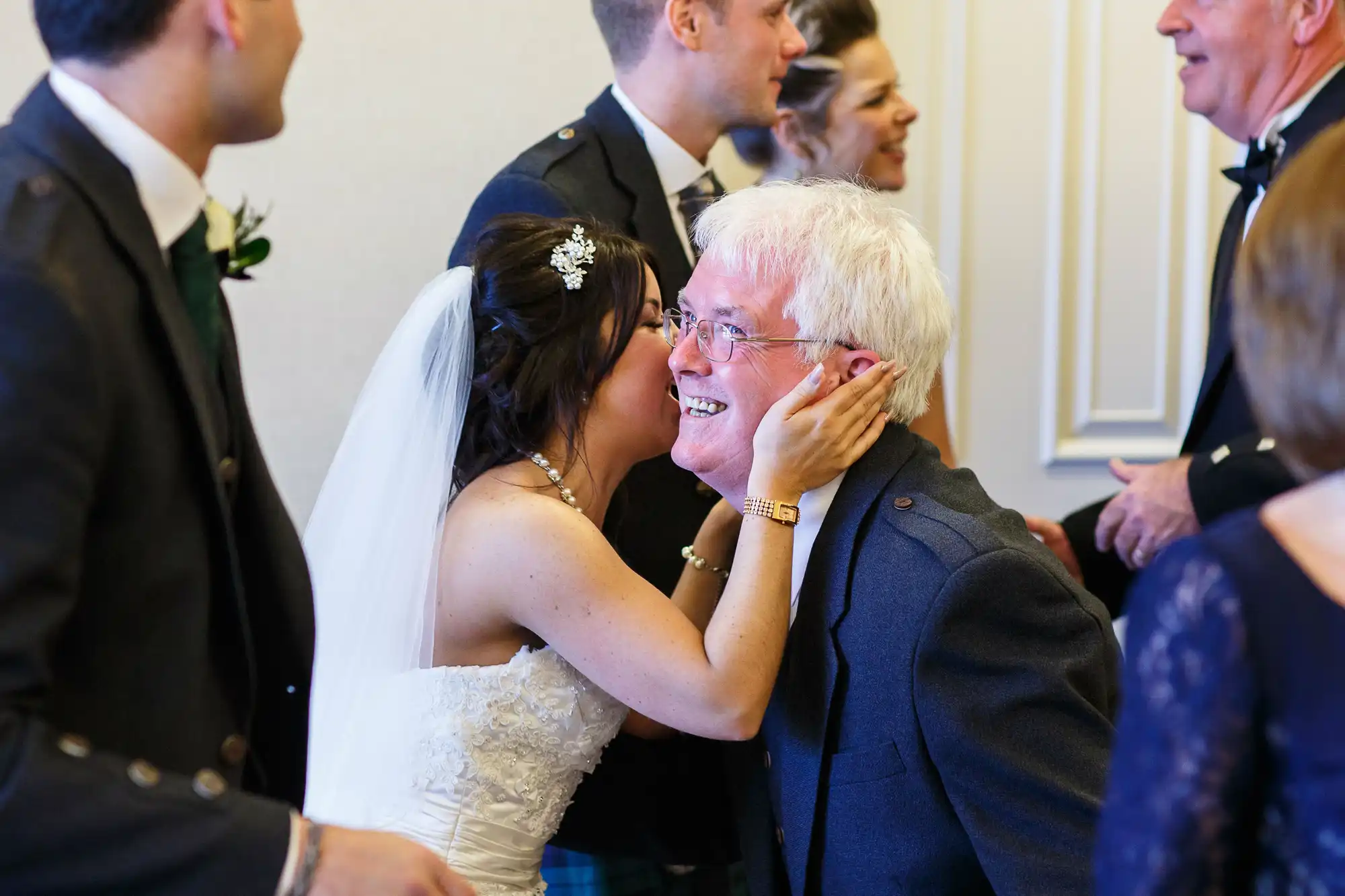 A bride in a white dress kisses a smiling older man on the cheek in a crowded room at a wedding celebration.