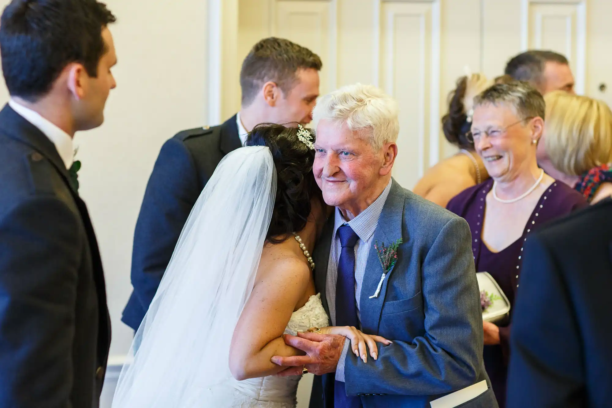 An elderly man in a blue suit embraces a smiling bride at a wedding ceremony, surrounded by guests.