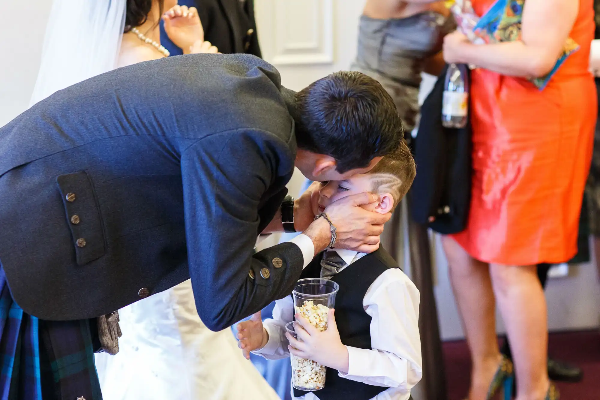 A man in a suit kisses a young boy on the forehead at a formal gathering, while the boy holds a cup of popcorn. people in the background are conversing.