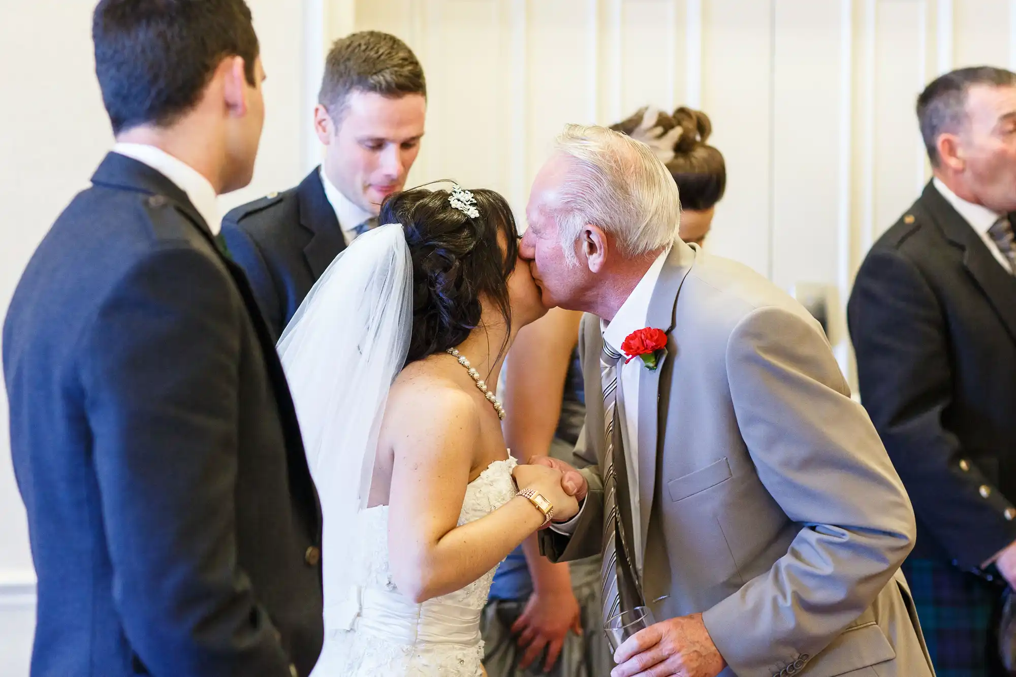 Elderly man kissing the bride on the cheek at a wedding reception, surrounded by guests in formal attire.