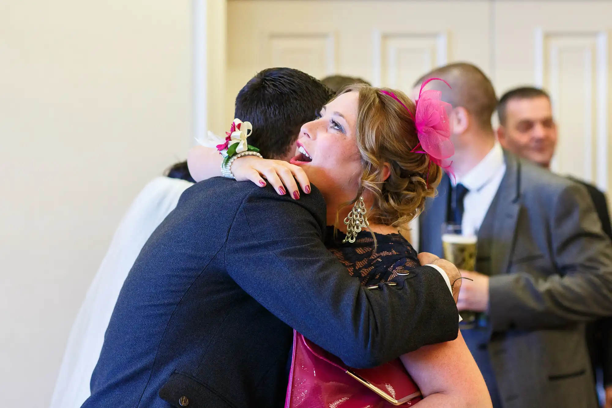 A woman in a pink dress and fascinator hugs a man in a dark suit at a social event, with another man smiling in the background.