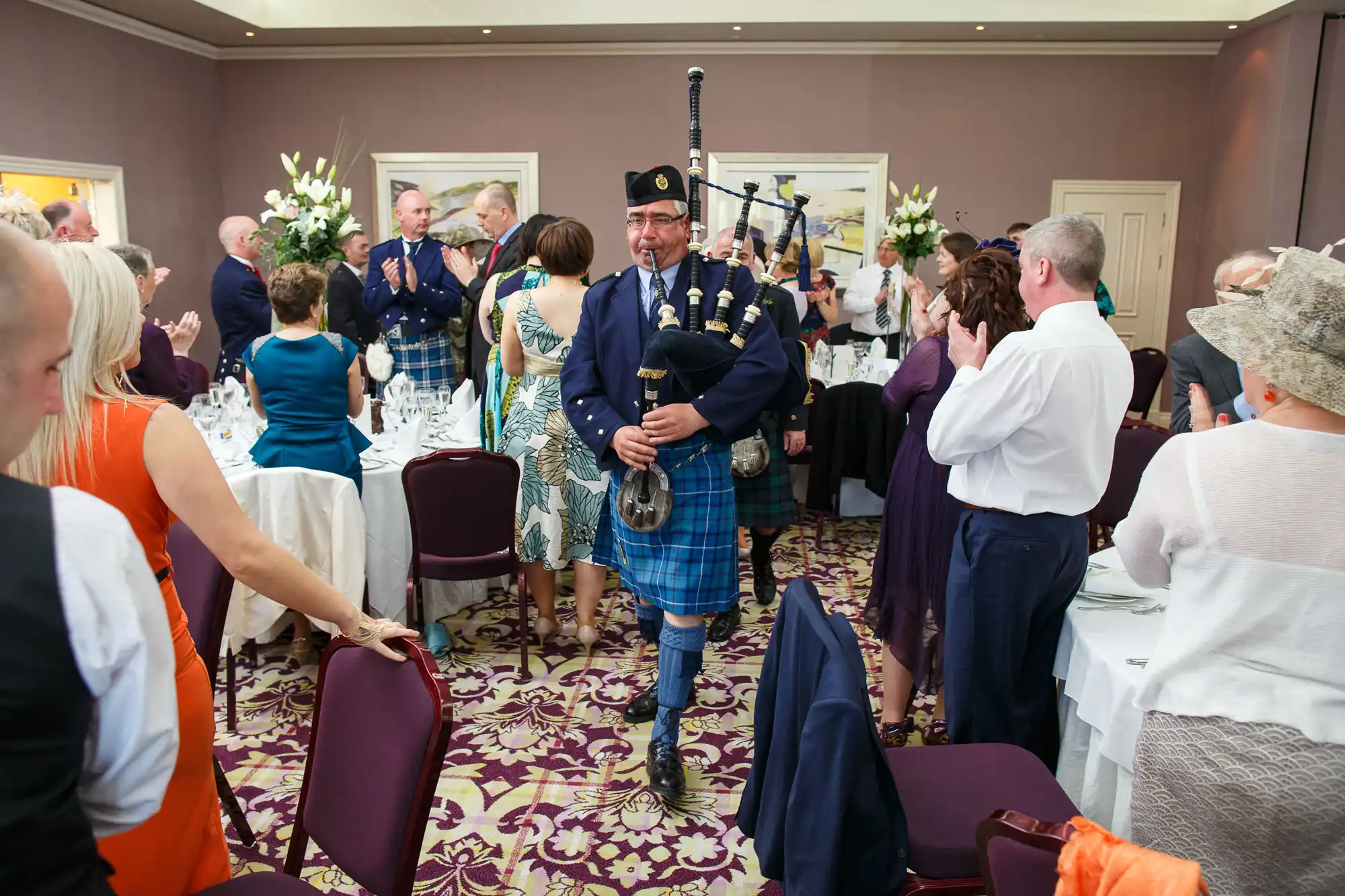 A bagpiper in traditional scottish attire performing at a wedding reception with guests clapping and observing.