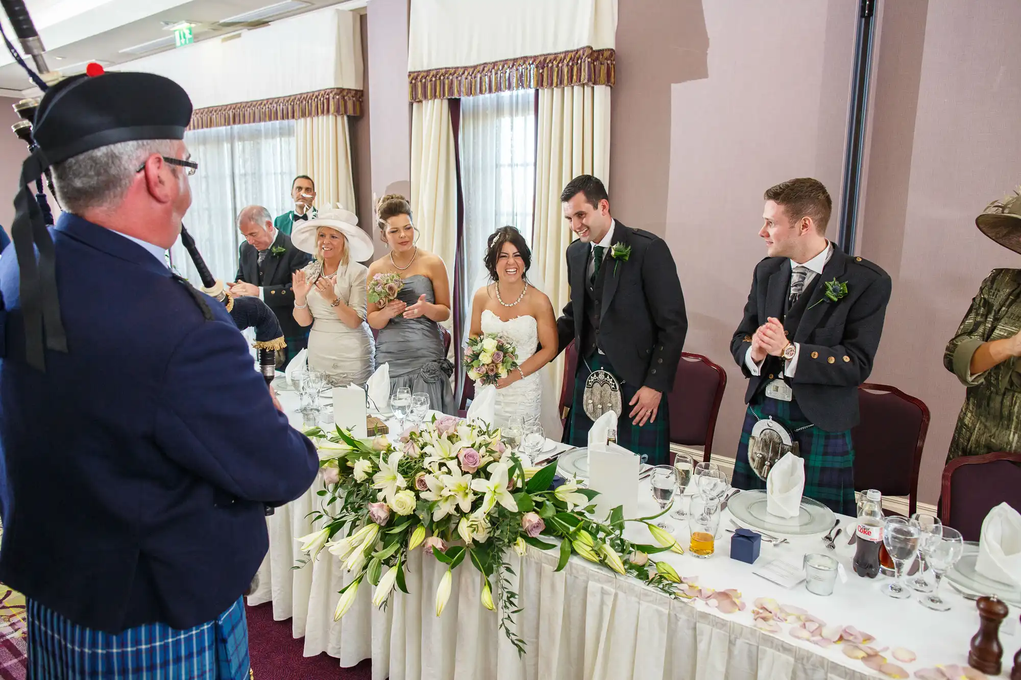 A wedding reception scene with guests smiling at a man in a kilt speaking at the head table, where a bride and groom stand beside floral arrangements.