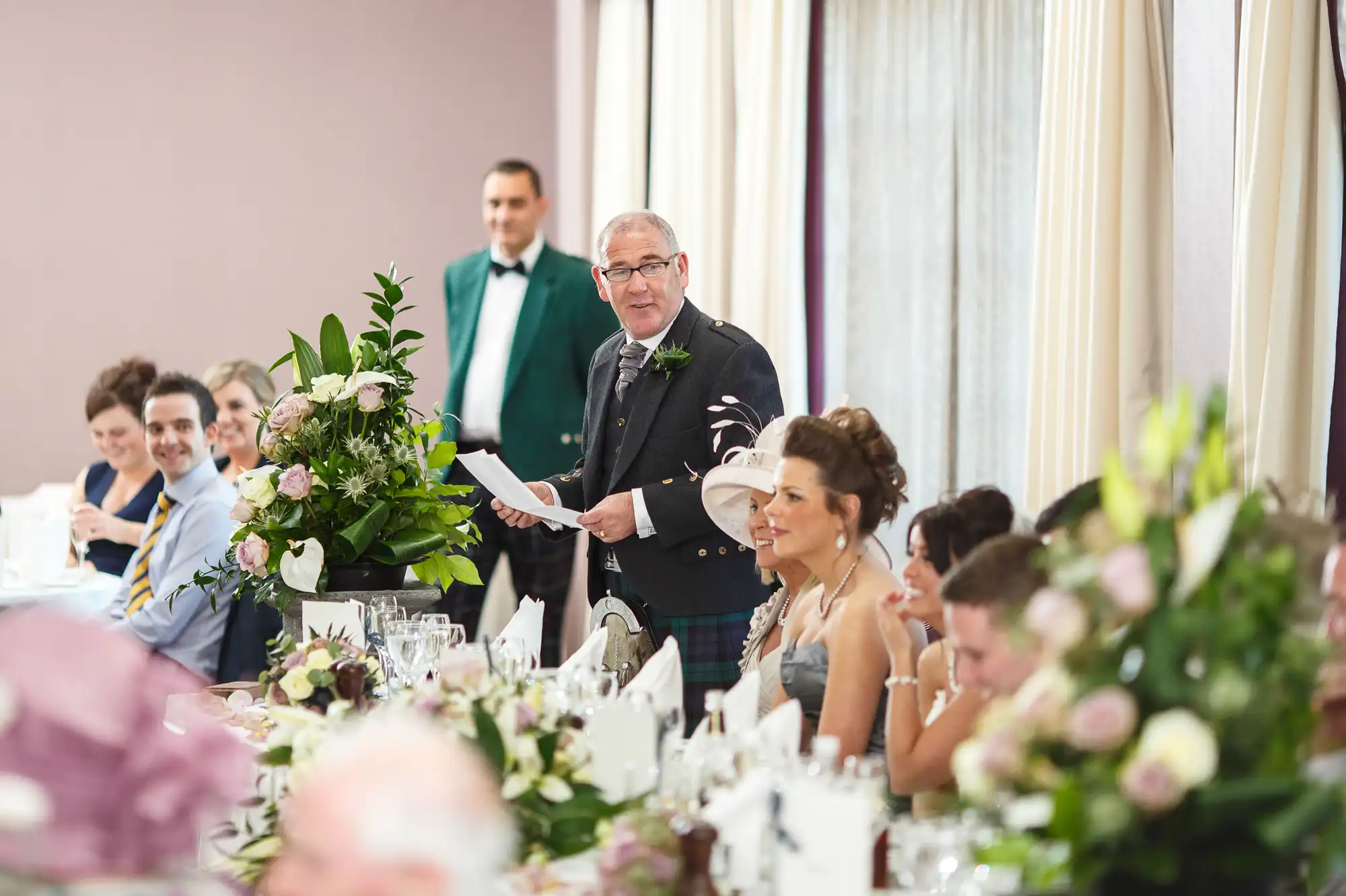 A man gives a speech at a wedding reception, holding a paper and a bouquet, with guests seated at tables, listening attentively.