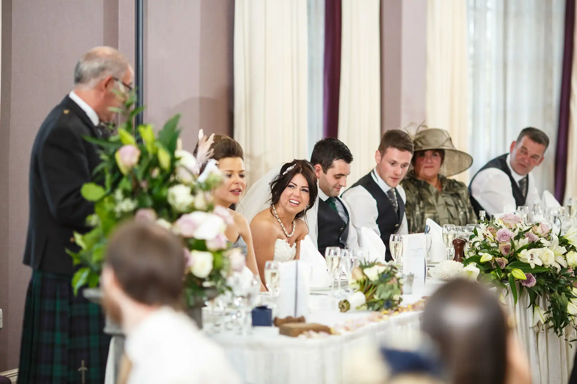 A joyful bride at a wedding reception table laughs with guests, some wearing traditional scottish attire, in an elegantly decorated room.