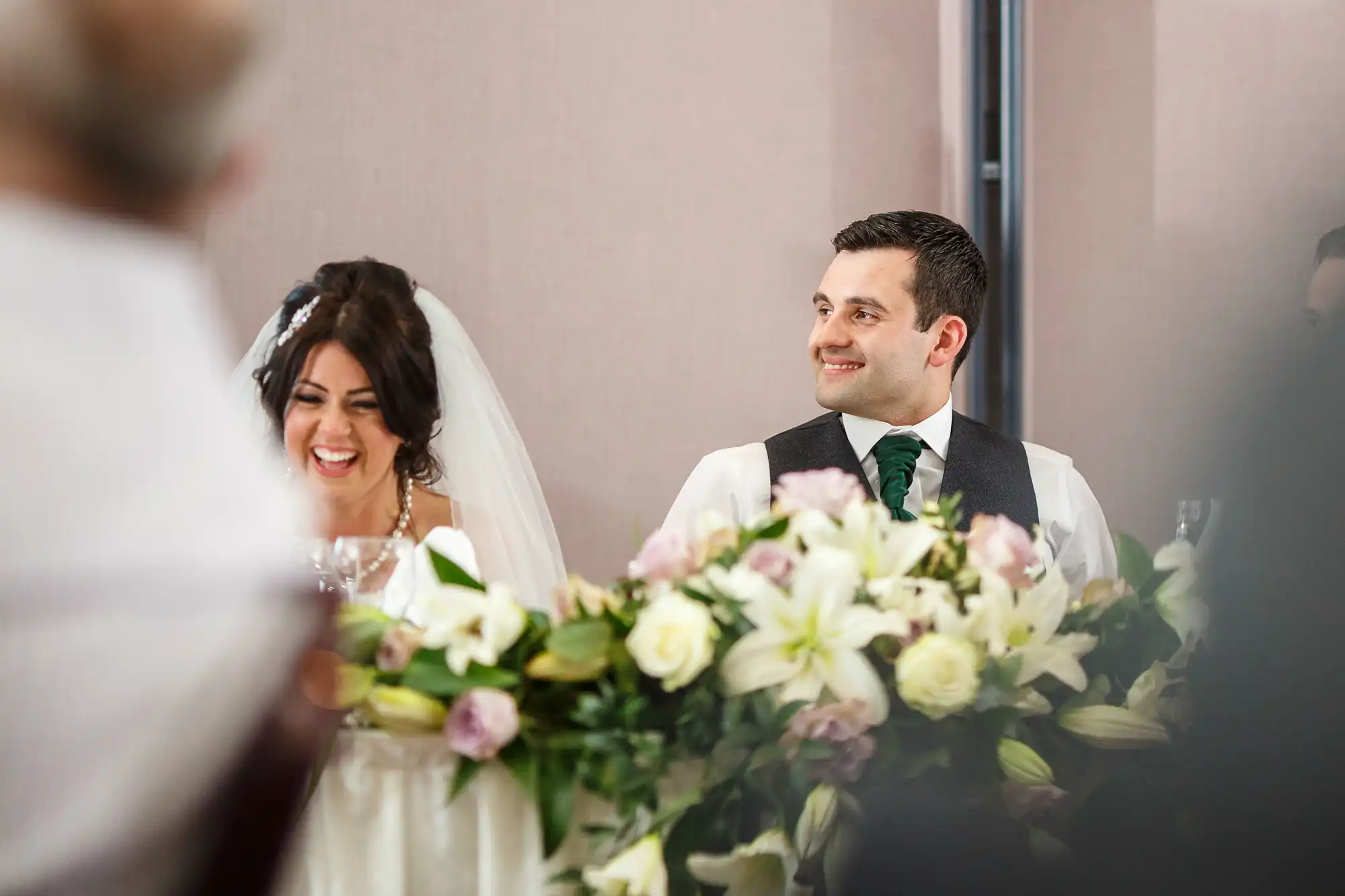 A bride and groom smiling at their wedding, with the groom looking at the bride as she laughs joyfully, surrounded by flowers.