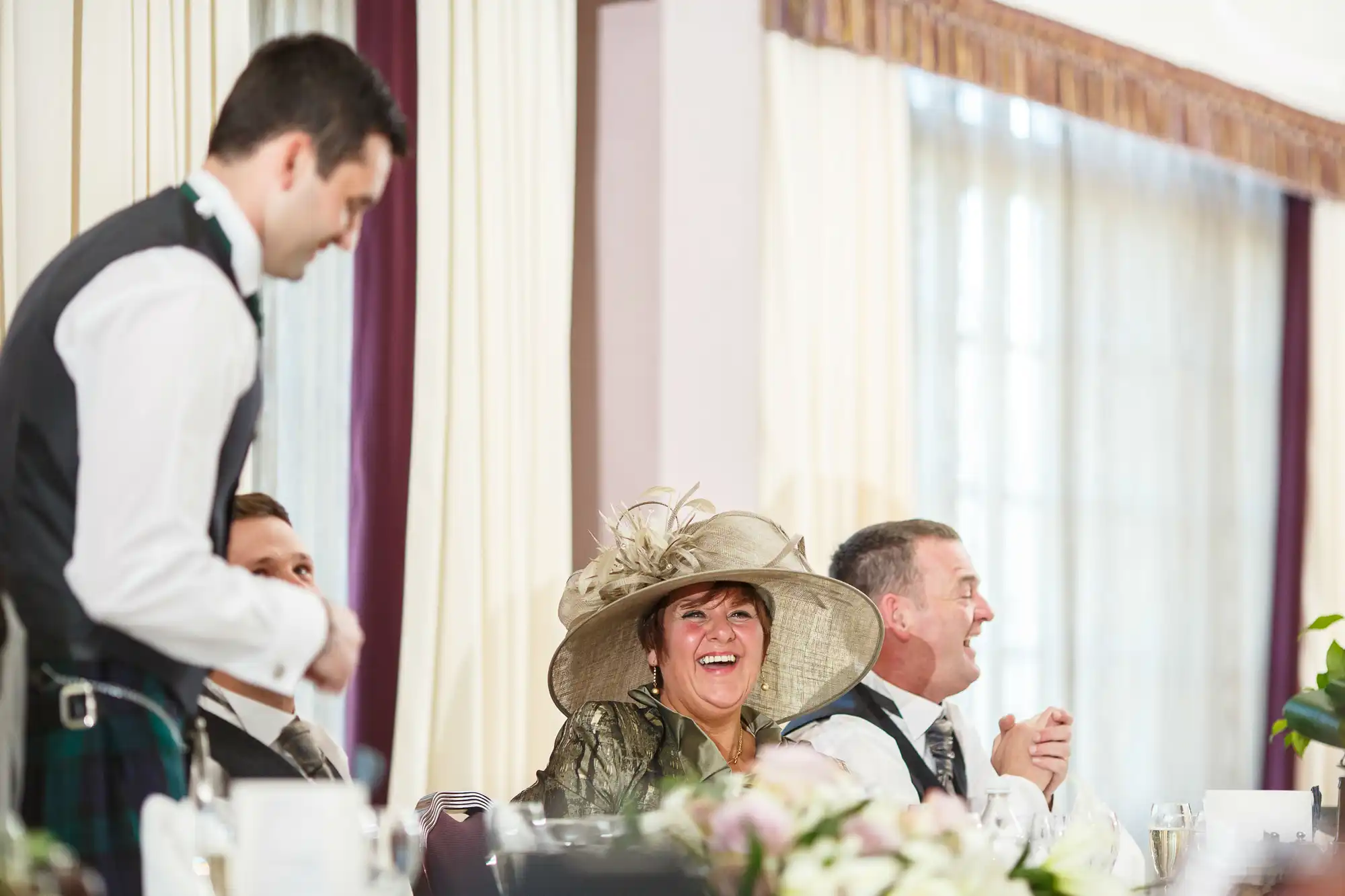 A woman in a large hat laughs joyfully at a formal dining event while a waiter serves and men in suits converse happily.