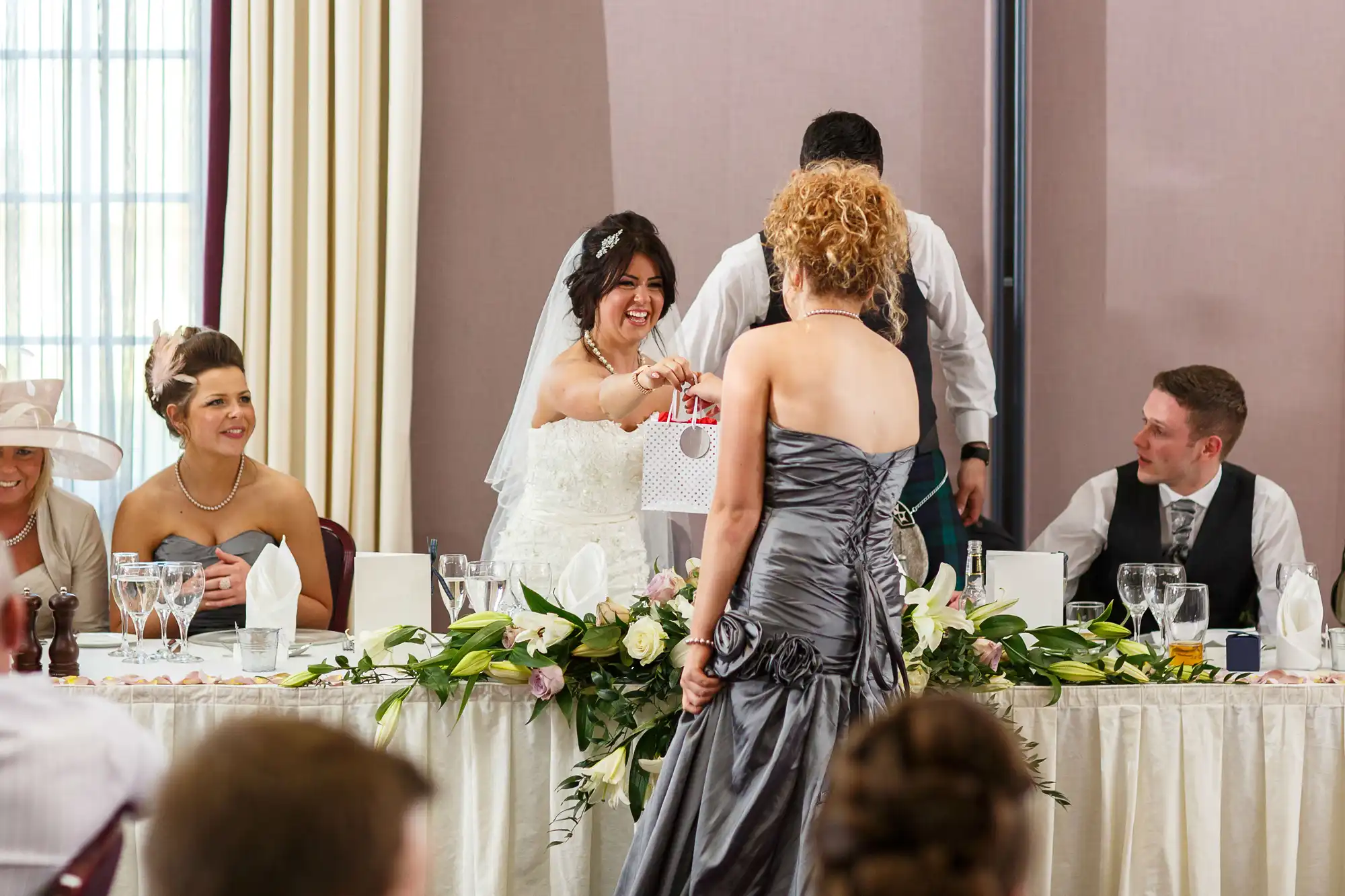 A bride in a white gown laughing and dancing with a guest at a wedding reception table surrounded by smiling guests.