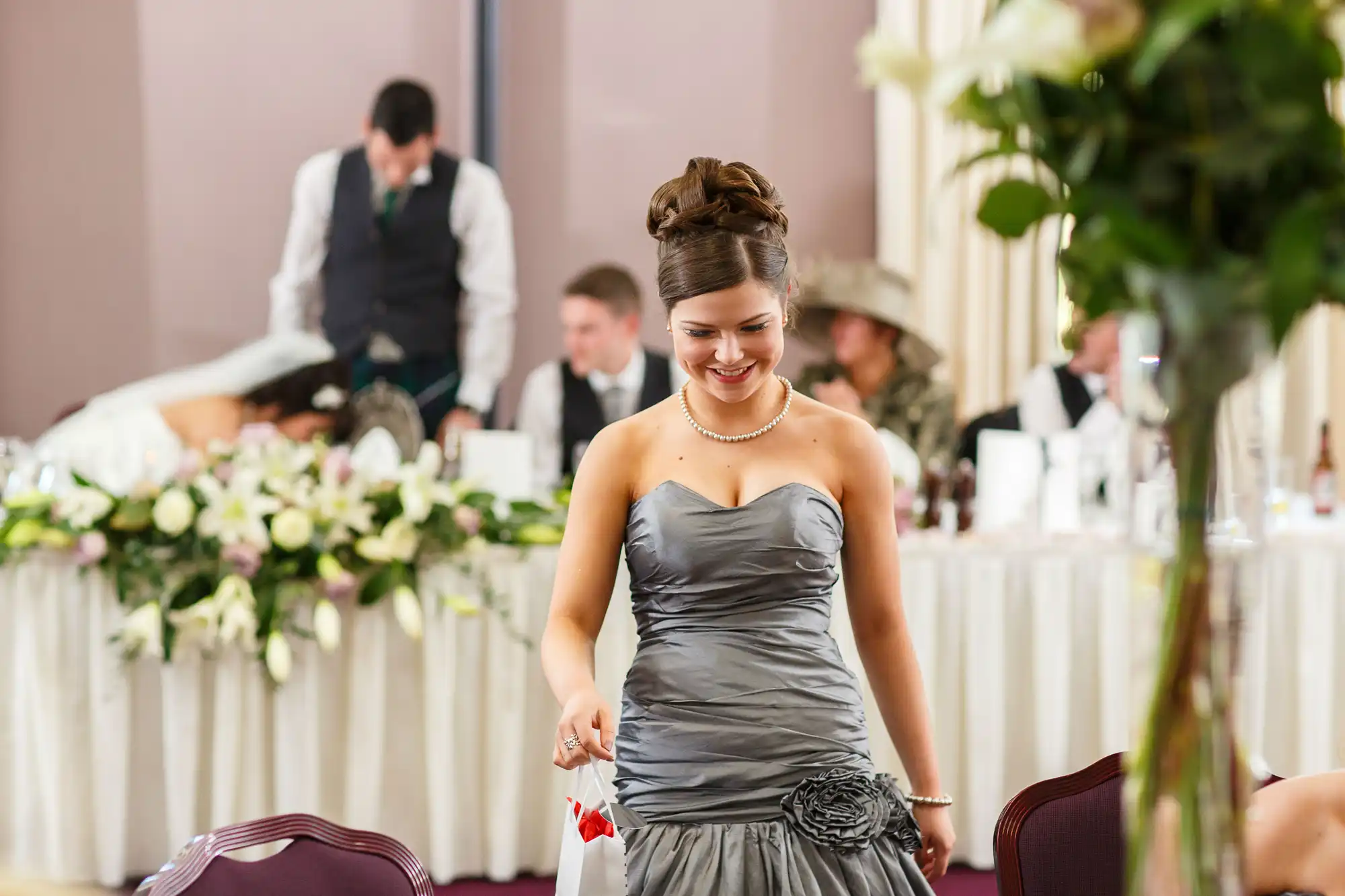 A woman in a gray dress smiles while walking at a wedding reception, with guests and tables in the background.