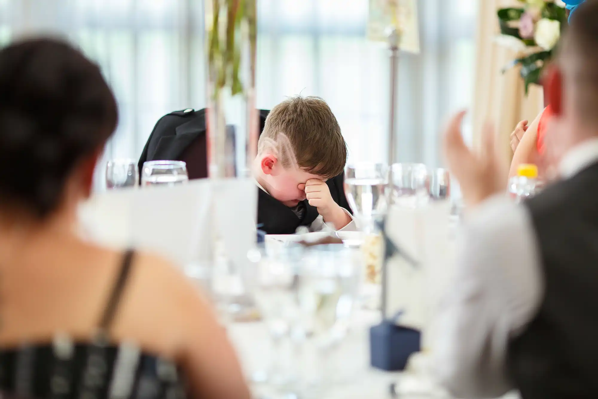 A young boy resting his head on his hand, looking bored or tired at a formal dining event. guests visible in the foreground.