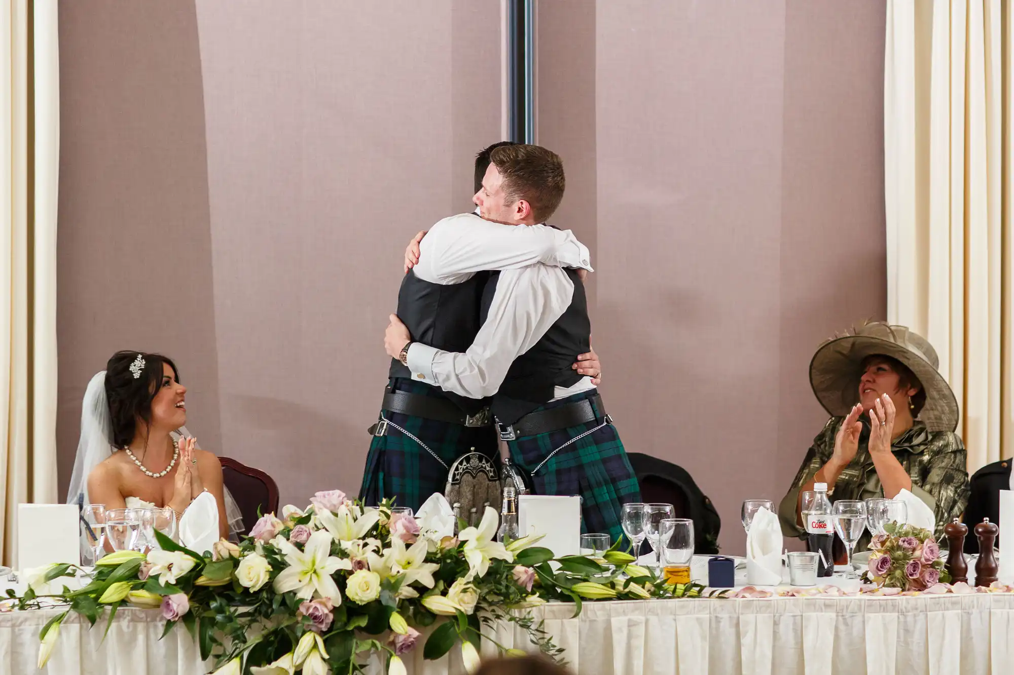 Two men in formal attire, one wearing a kilt, embrace at a wedding reception as seated guests look on smiling.