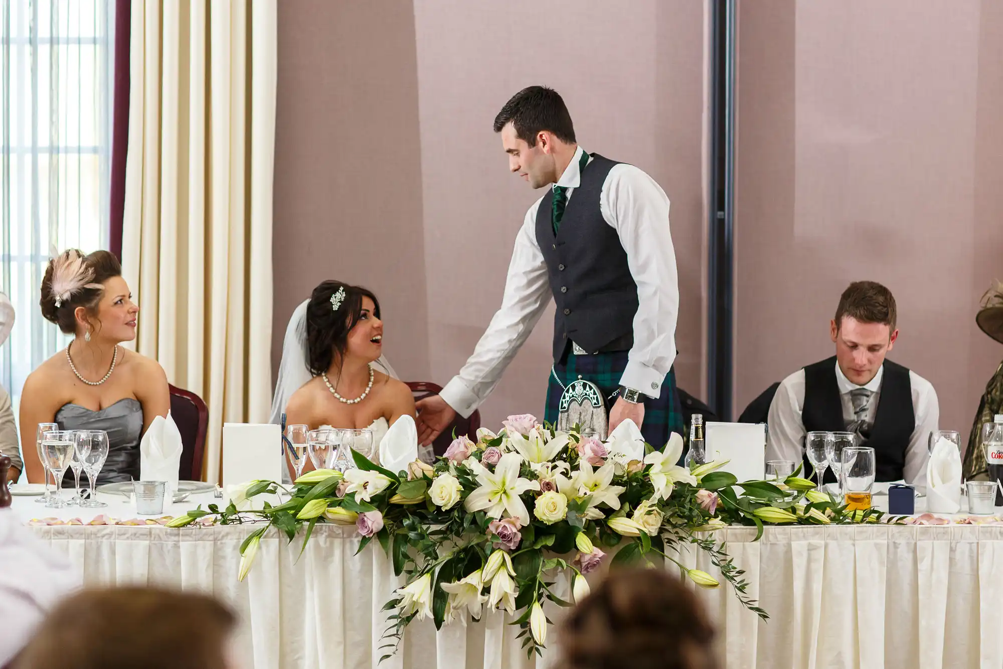 A groom in a kilt holding hands with his bride at a wedding reception table, surrounded by guests and floral decorations.