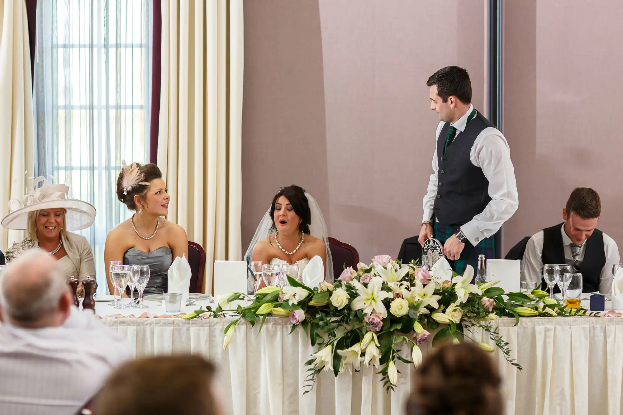 A man stands speaking at a wedding reception table, facing a group of seated guests who are visibly engaged and reacting with various expressions.