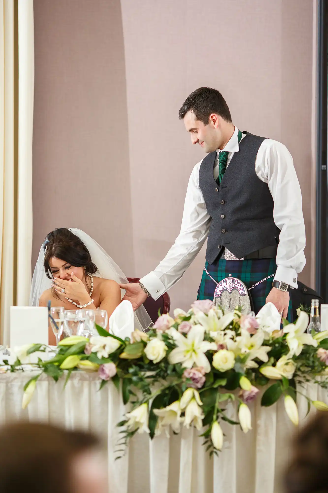 Groom in a kilt comforting bride sitting at wedding table, surrounded by flowers and white table settings.