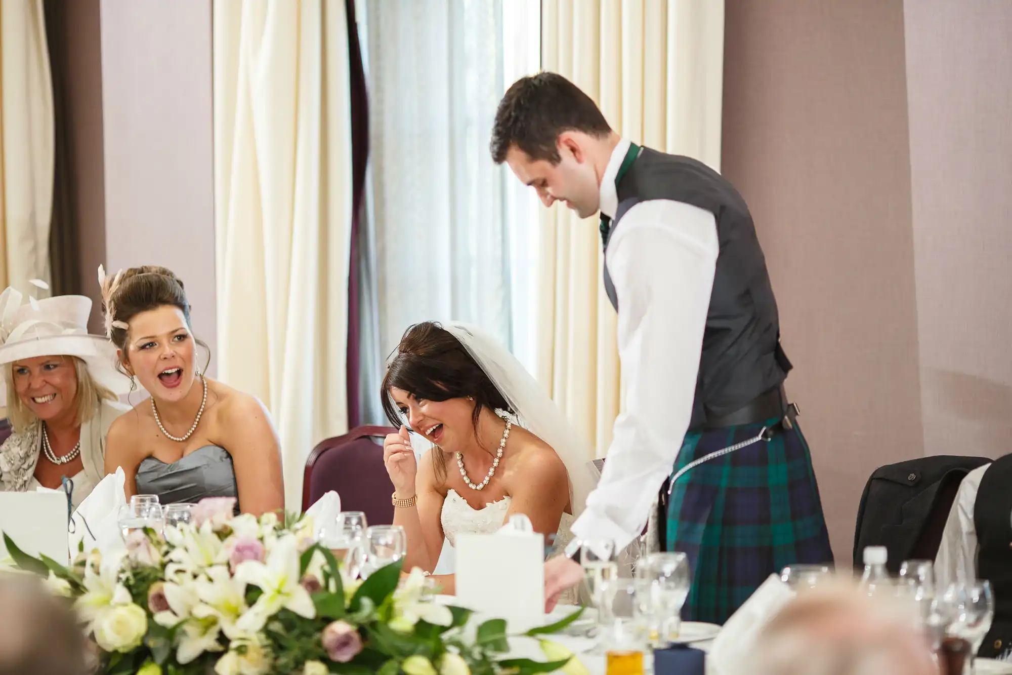 A bride and groom at a wedding reception table, laughing with guests, the groom wearing a kilt.