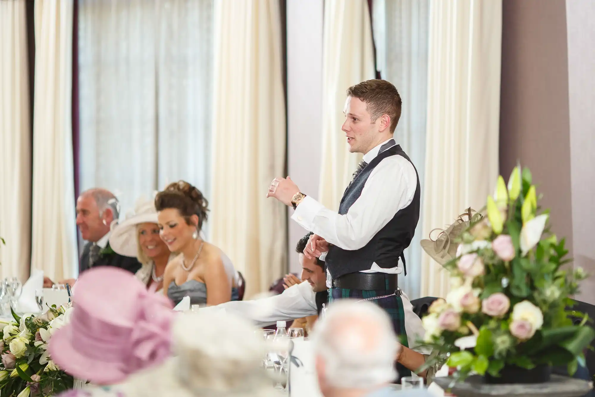 A man giving a speech at a wedding reception, gesturing with his right hand, with guests seated around tables, listening and smiling.