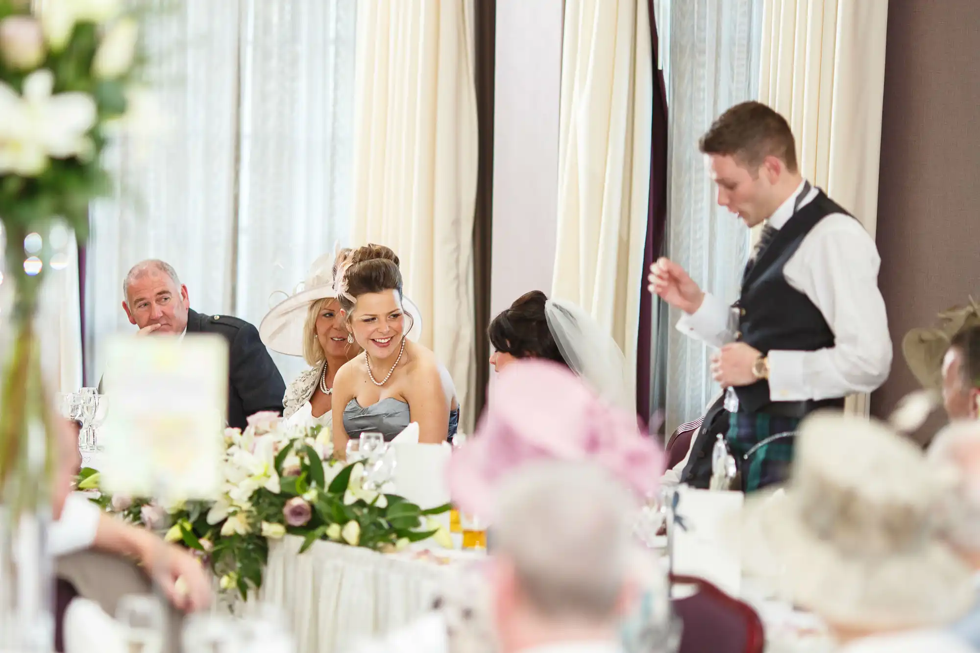 A joyful wedding reception with guests at tables; a man in a kilt is speaking as people listen and smile.