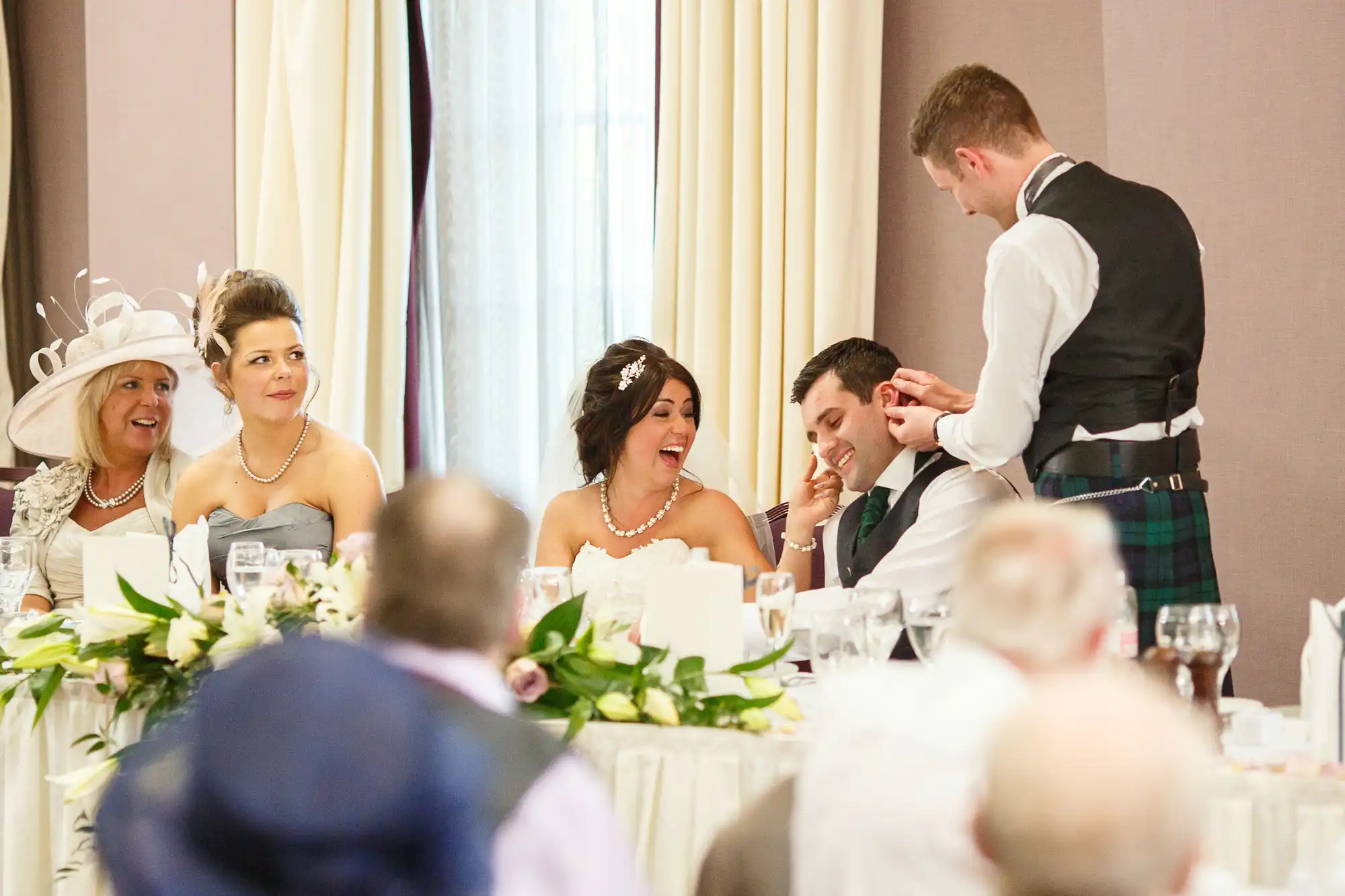 A wedding reception scene with guests dressed in formal wear, including kilts, laughing and interacting around a table.