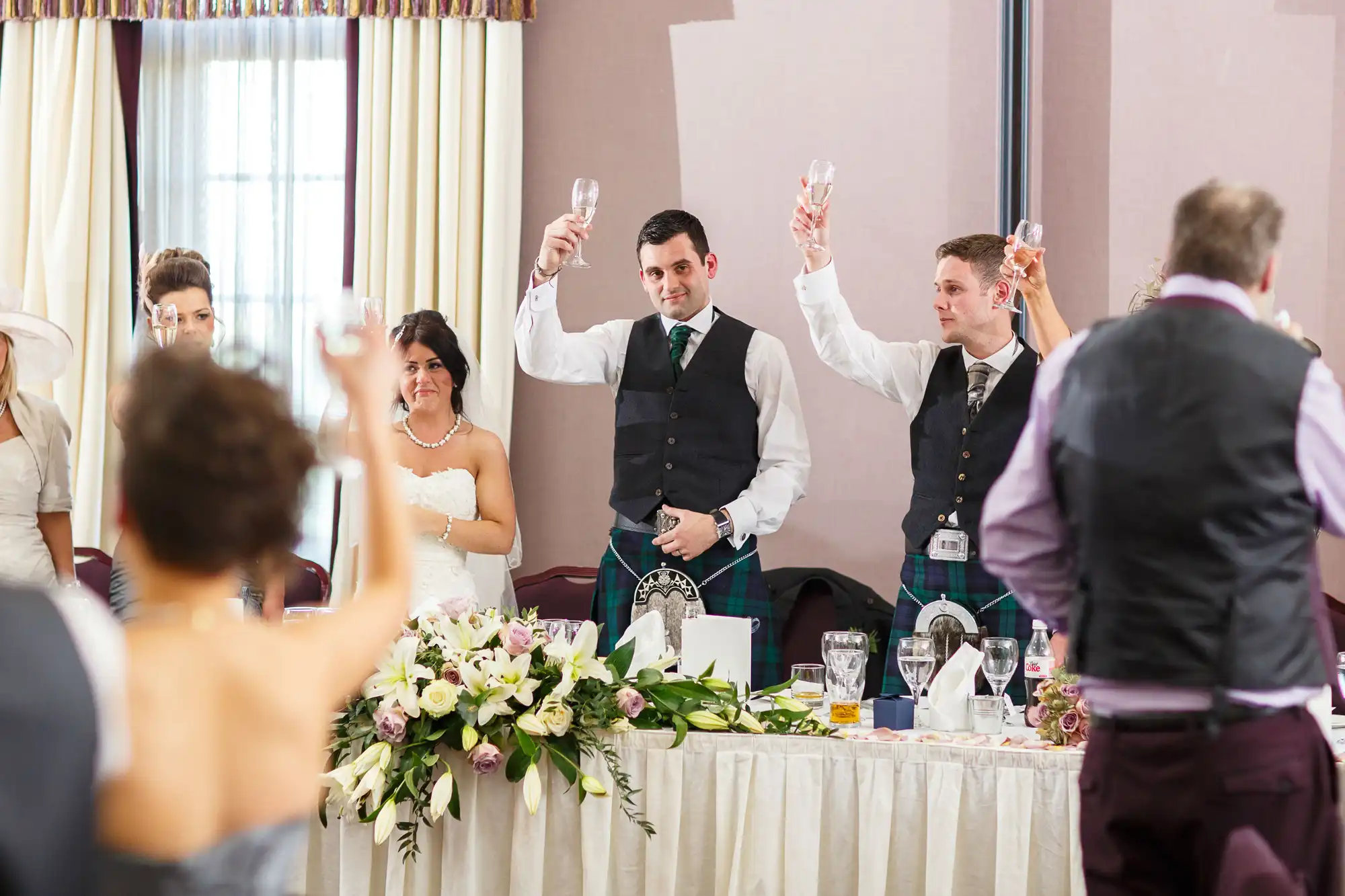 A bride and groom in traditional scottish attire raise their glasses for a toast at their wedding reception, surrounded by guests.