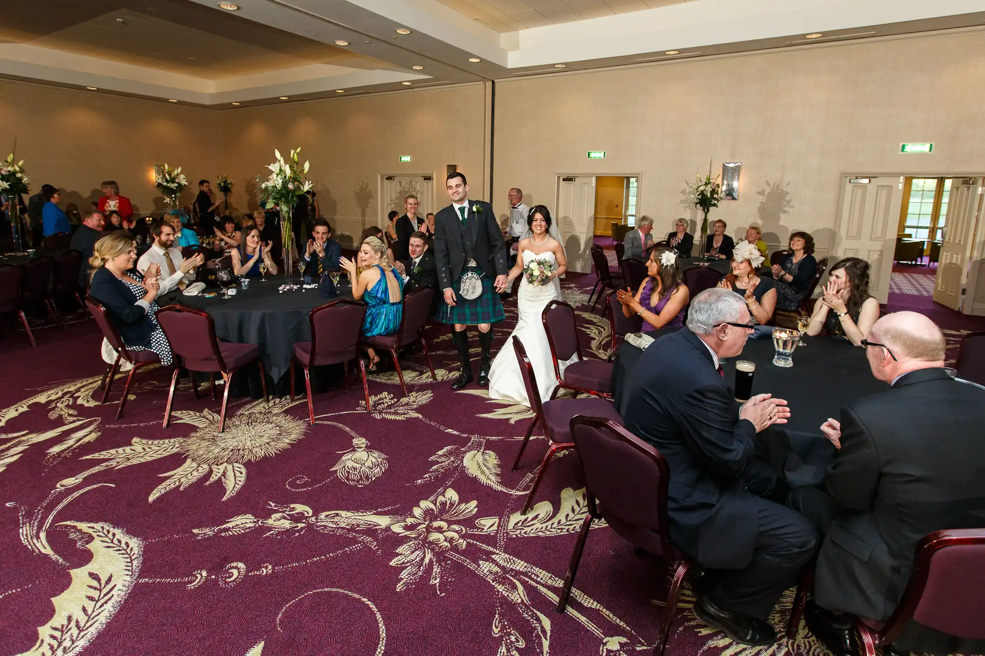 A bride and groom walking through a banquet hall as guests seated at tables applaud them.