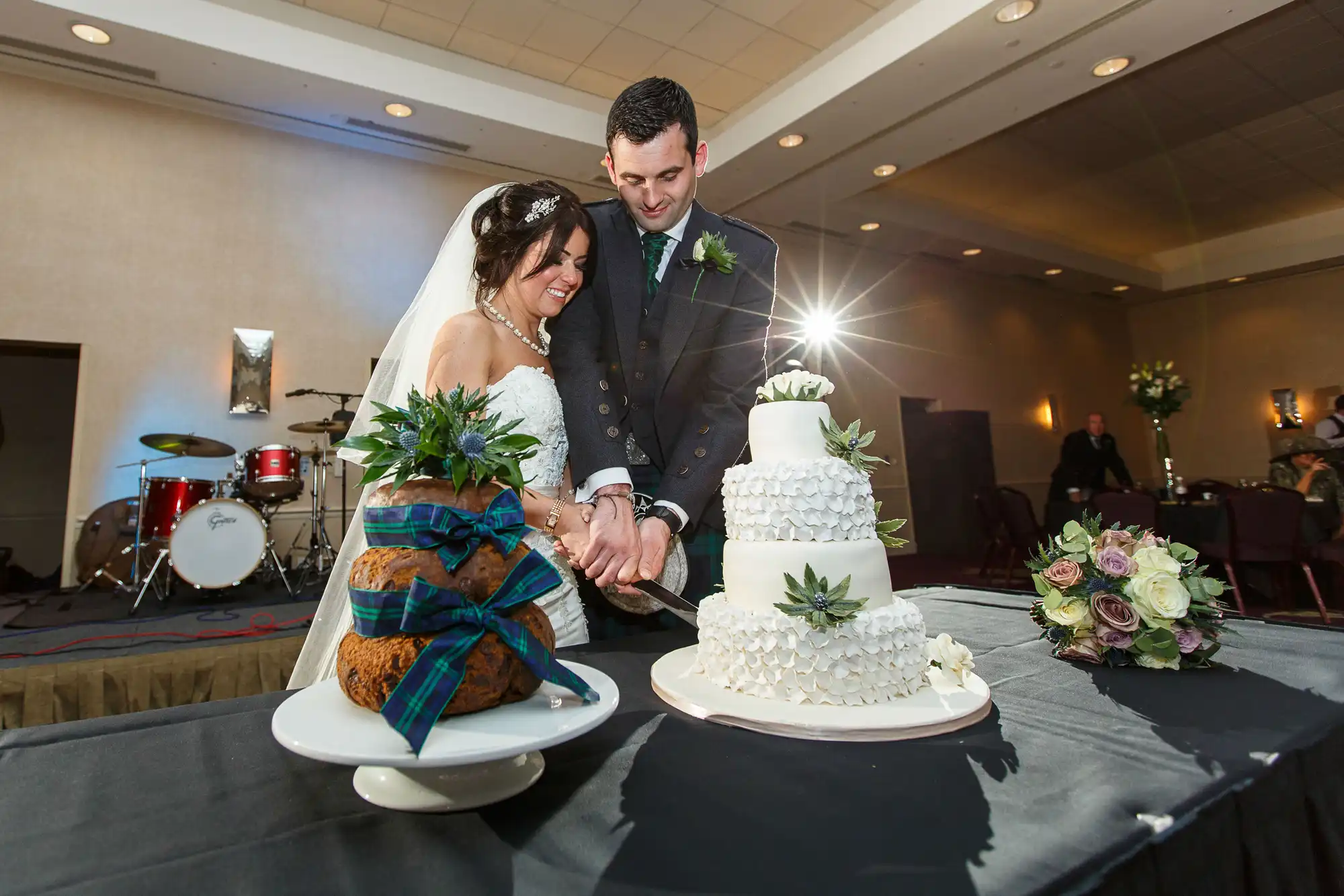 A bride and groom cutting their wedding cake, smiling, with a band setup in the background.