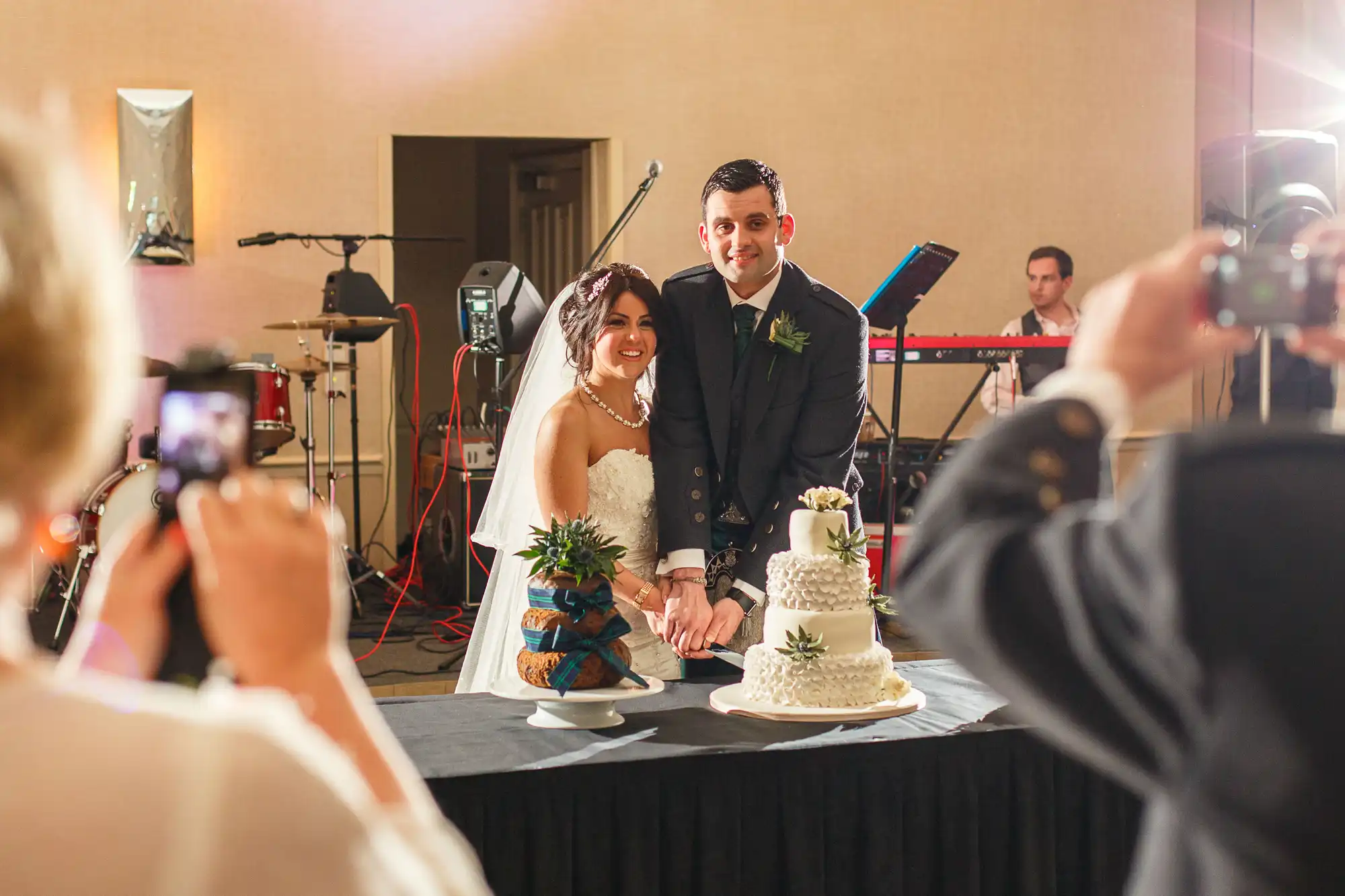 A bride and groom smiling beside their wedding cake, surrounded by guests capturing the moment with cameras.