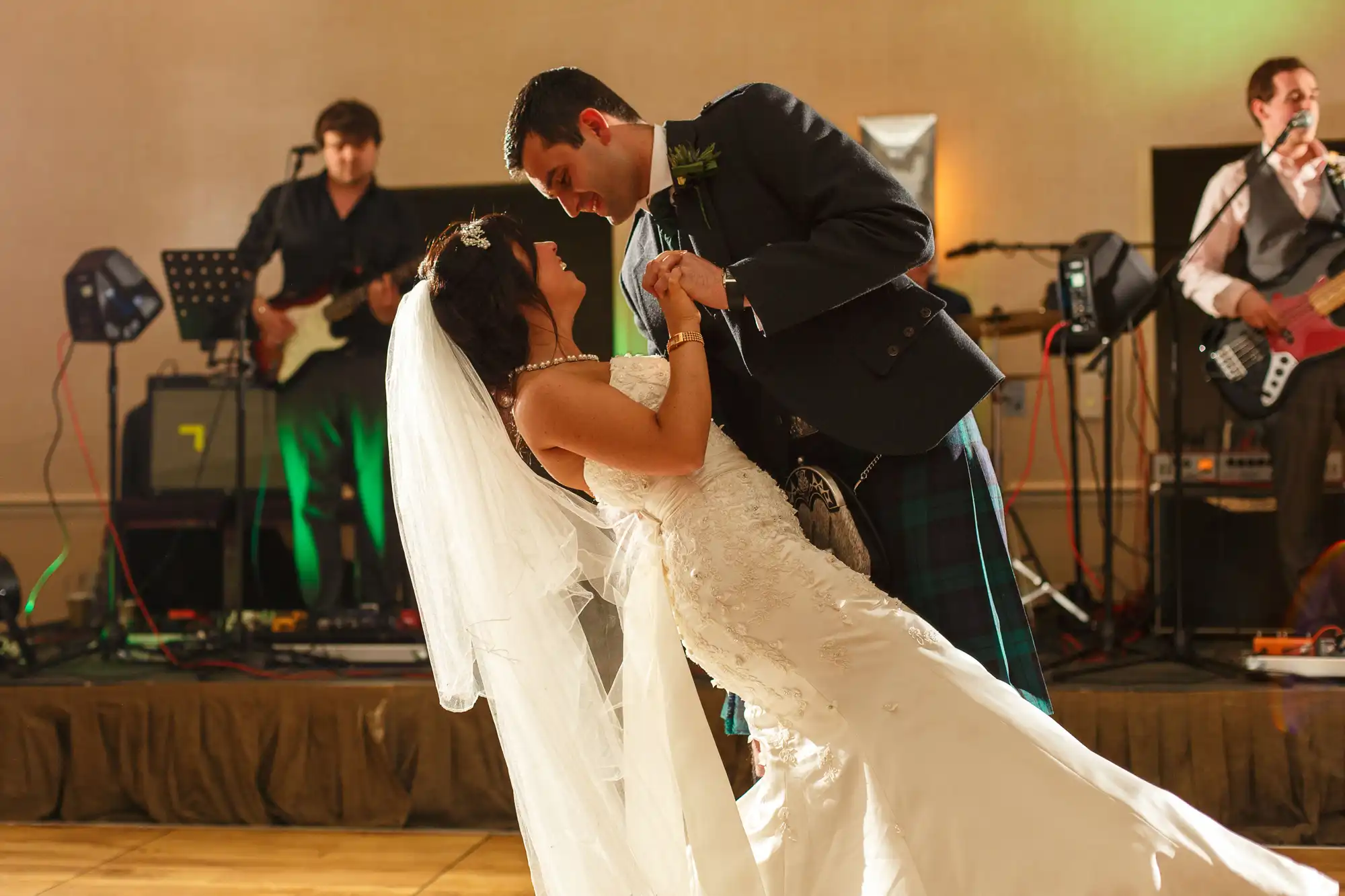 A bride and groom share a dance in a ballroom with a live band performing in the background. the groom is in a kilt and the bride is in a long white dress.