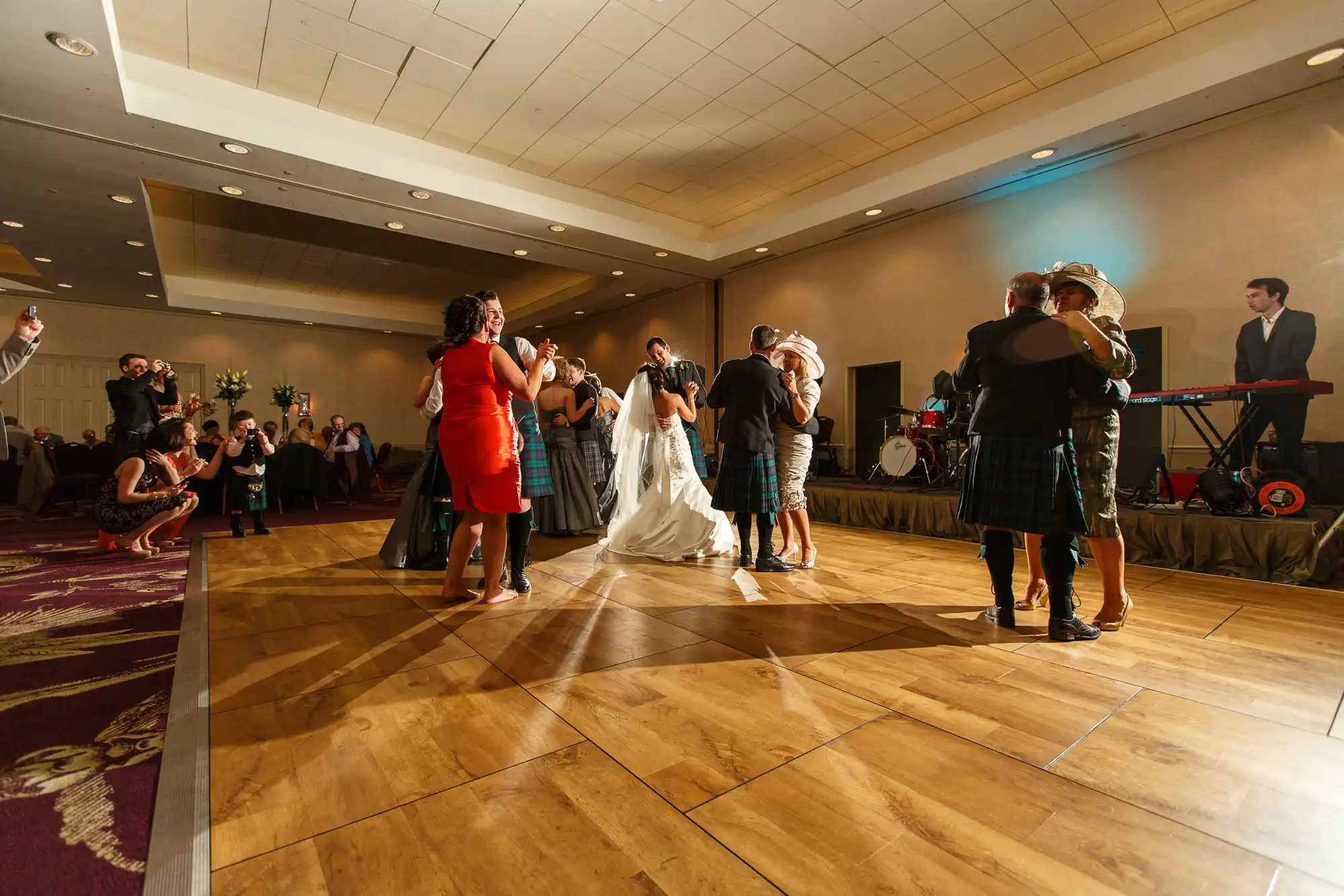 Wedding guests dance on a wooden floor in a banquet hall, with a bride in white and several guests wearing kilts, as onlookers watch from the sidelines.