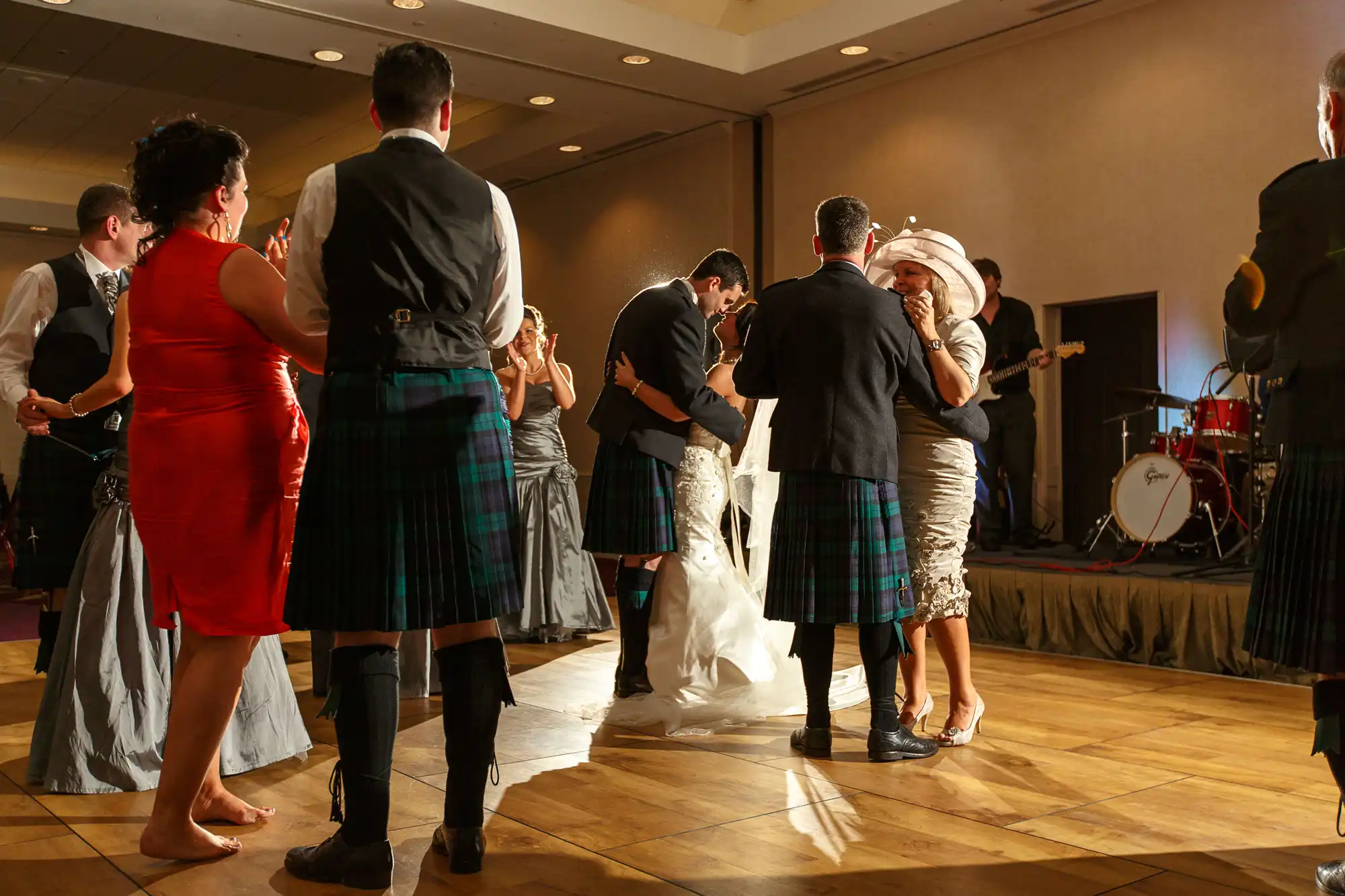 People in formal attire, including kilts, dance closely at a warmly lit indoor event with a live band in the background.
