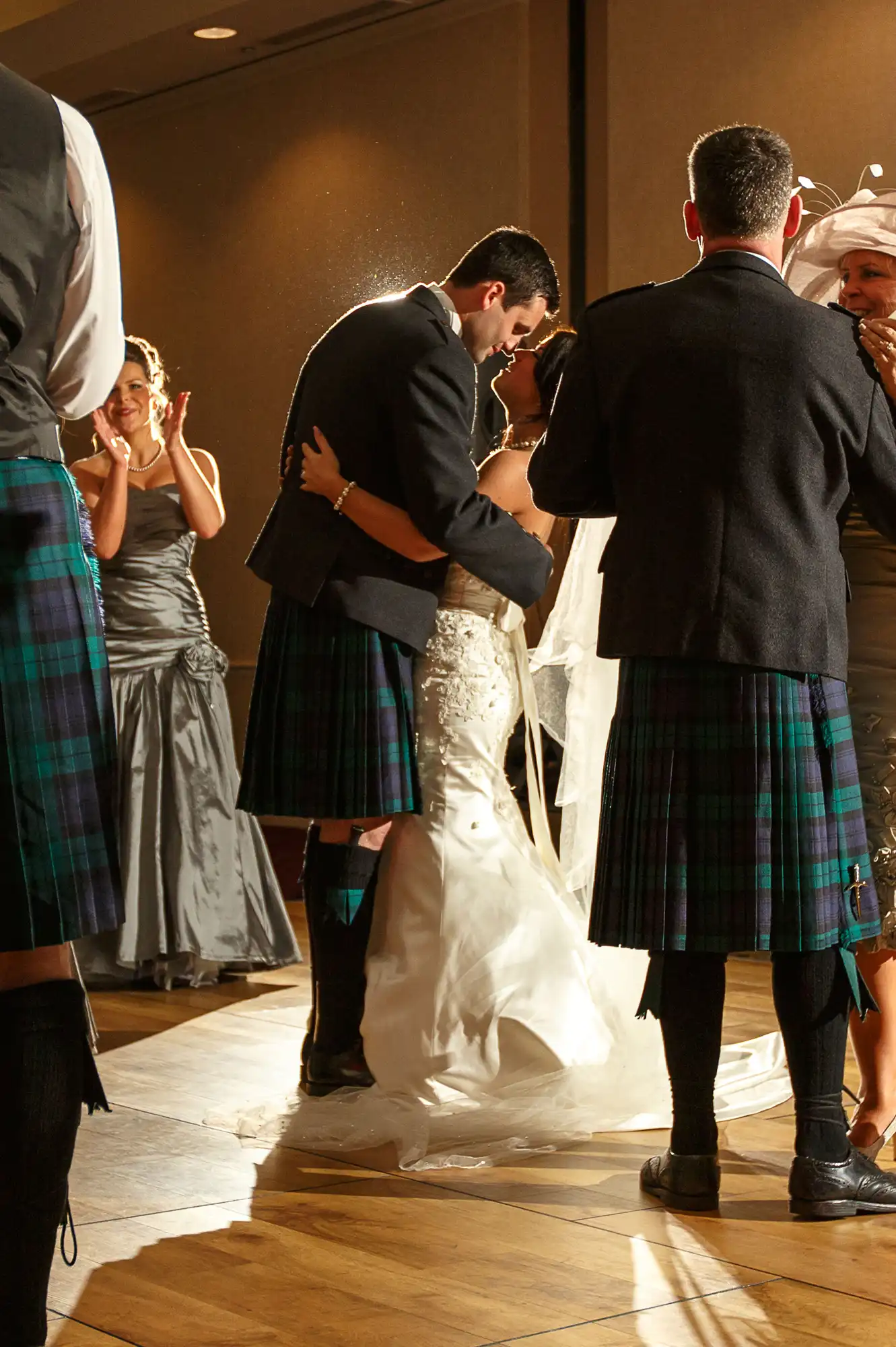 A couple in wedding attire, including kilts, share a kiss on a dance floor surrounded by applauding guests.