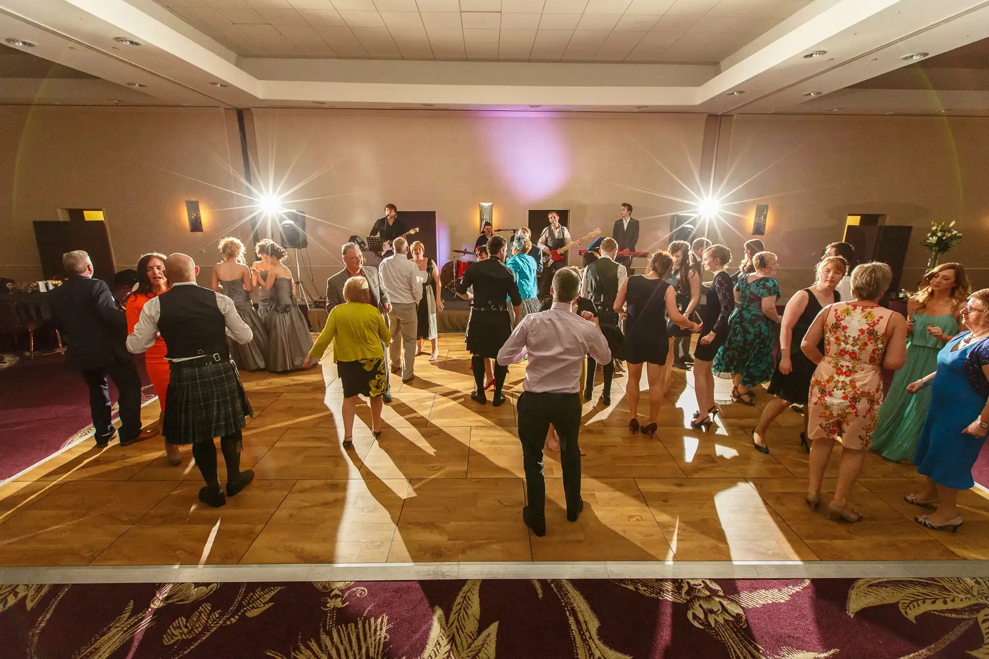 Guests dancing at a wedding reception in a ballroom with a live band playing in the background and colorful lighting.