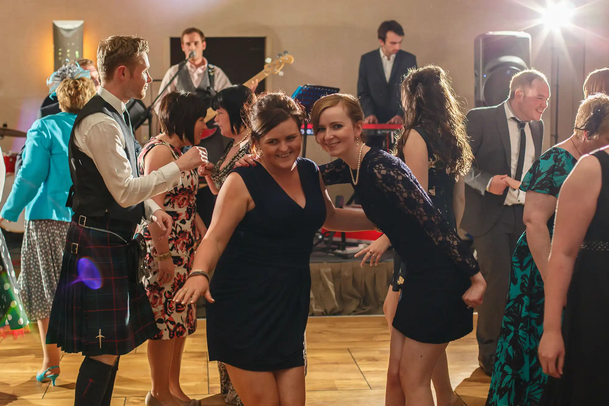 People dancing and smiling at a festive indoor event with a live band playing in the background. some men wear kilts and women are in dresses.