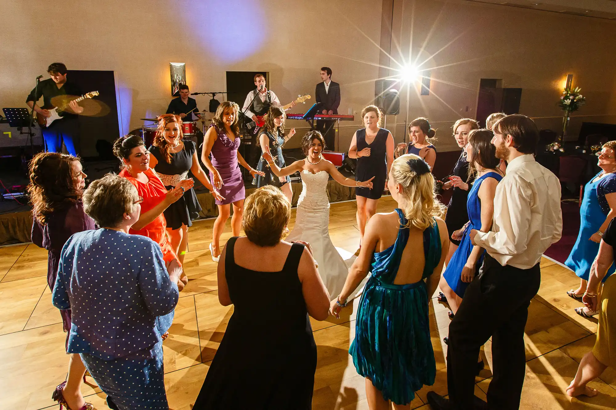 A lively wedding reception dance floor with guests dancing in a circle around a bride, a live band performing in the background.