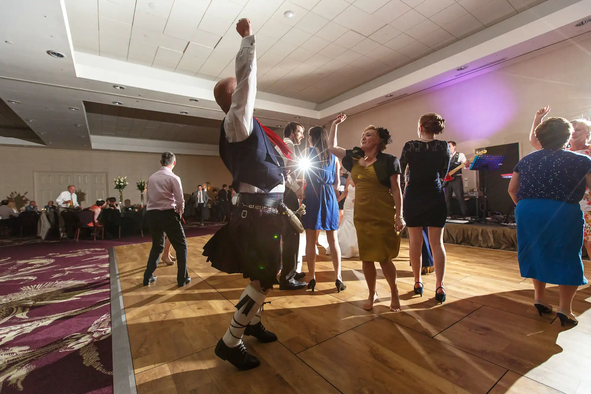 Guests dancing joyously at a wedding reception, with a man in a kilt raising his arm triumphantly under a bright light.