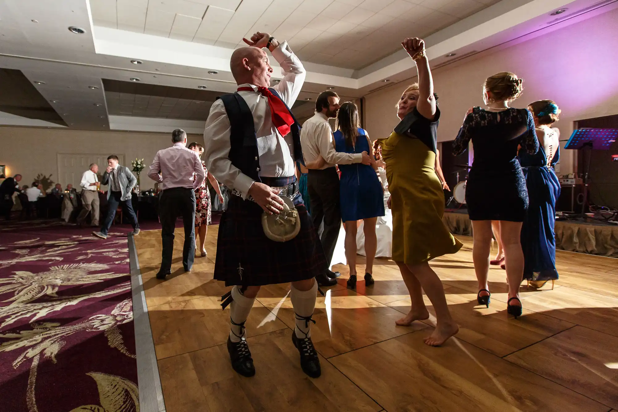 People dancing energetically at an indoor celebration, one man wearing a traditional kilt. brightly lit room with dancing couples and a colorful lighting setup in the background.