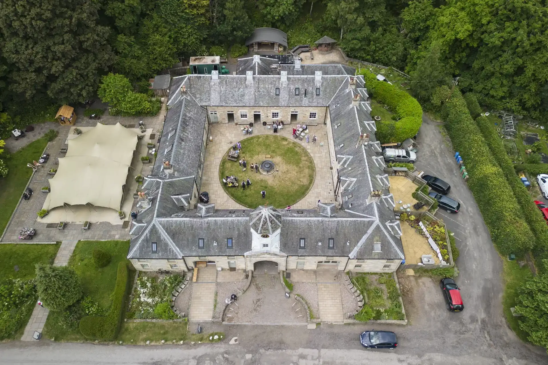 Aerial view of a large, historic stone building with a courtyard, surrounded by lush green trees, with several cars parked around.
