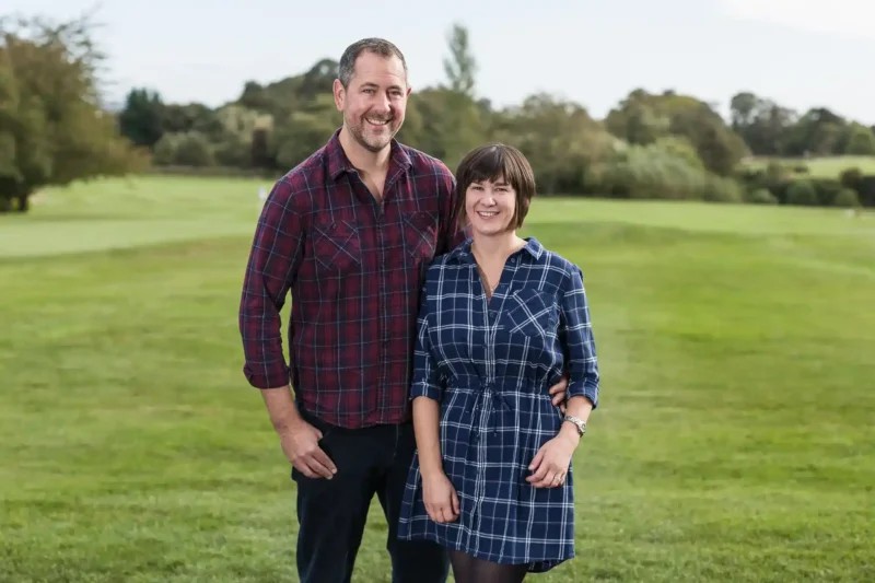 A man and a woman stand together on a grassy field, both smiling at the camera. Trees and shrubbery are visible in the background.