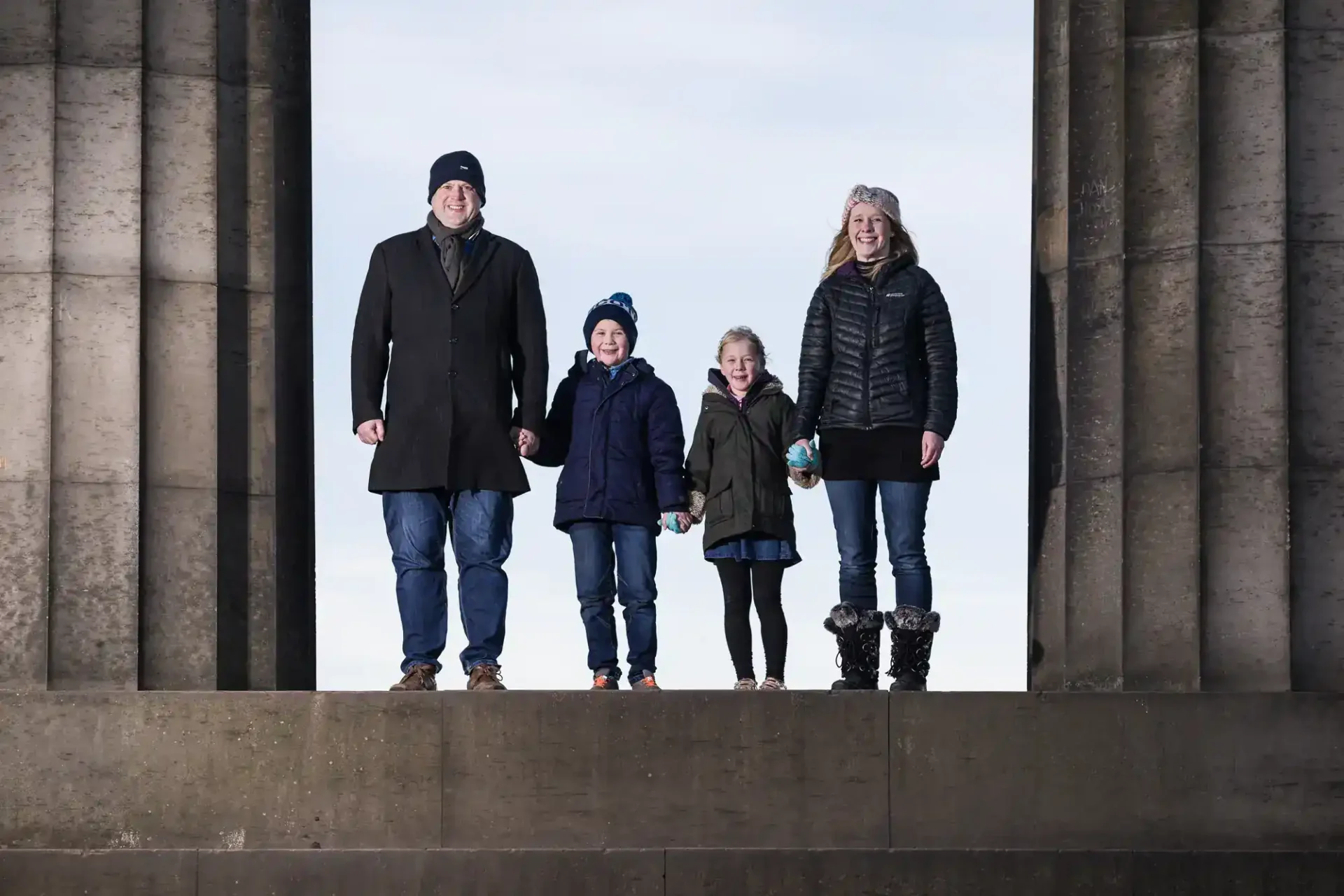 A man, woman, and two children stand together outdoors between large stone columns, all dressed in winter clothing.