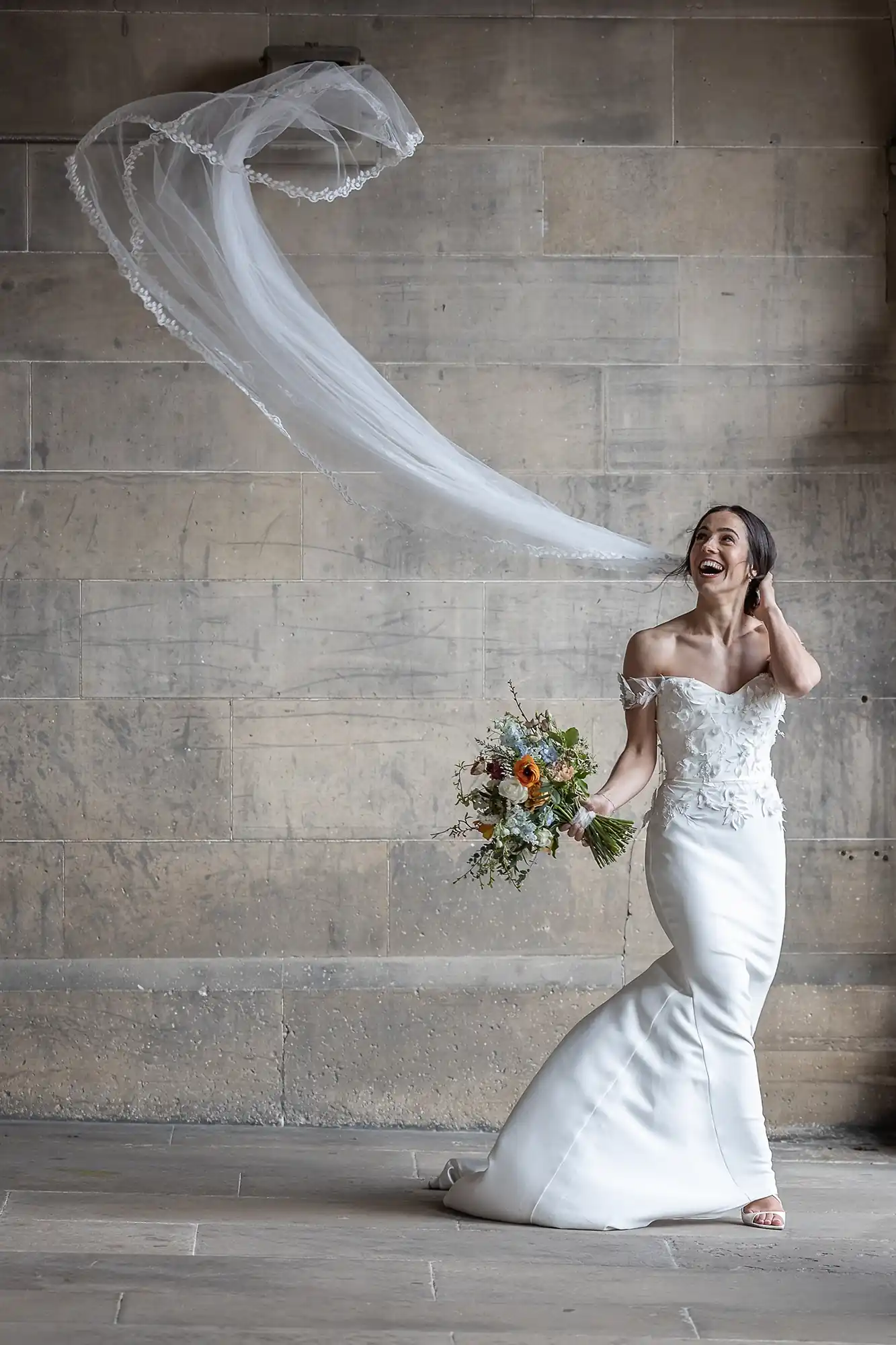 Bride in an off-shoulder gown holding a bouquet, laughing as her veil flows in the air against a stone wall backdrop, image by Fettes wedding photographer Edinburgh